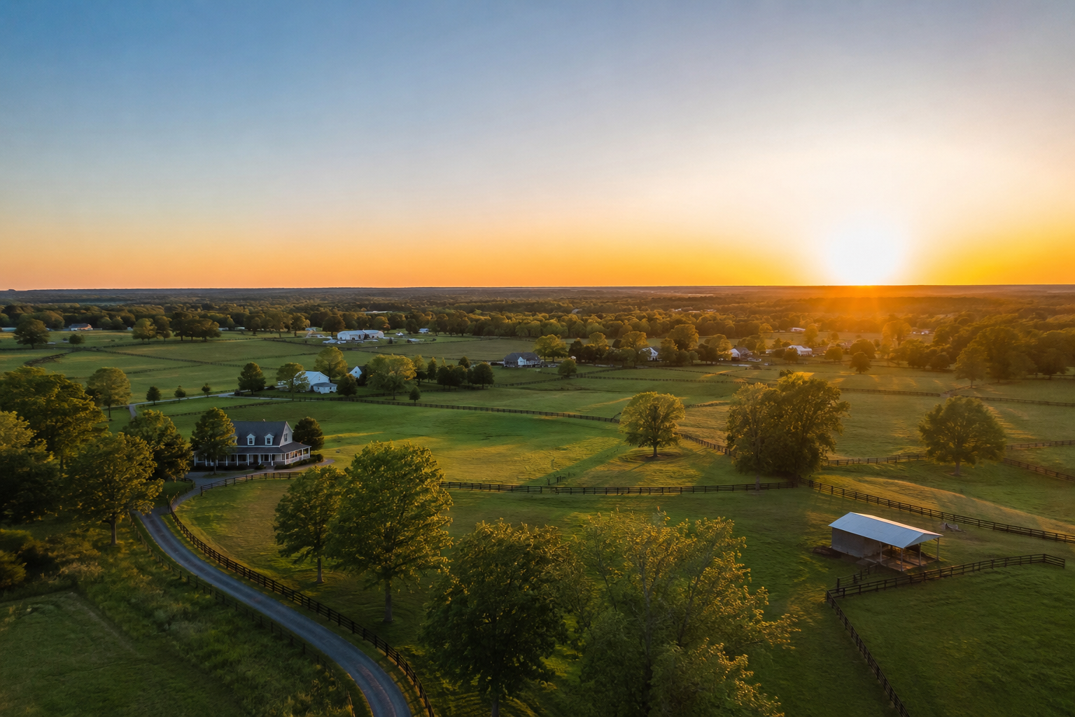A scenic rural landscape at sunset featuring a house, trees, green fields, winding roads, and a small barn surrounded by fences.