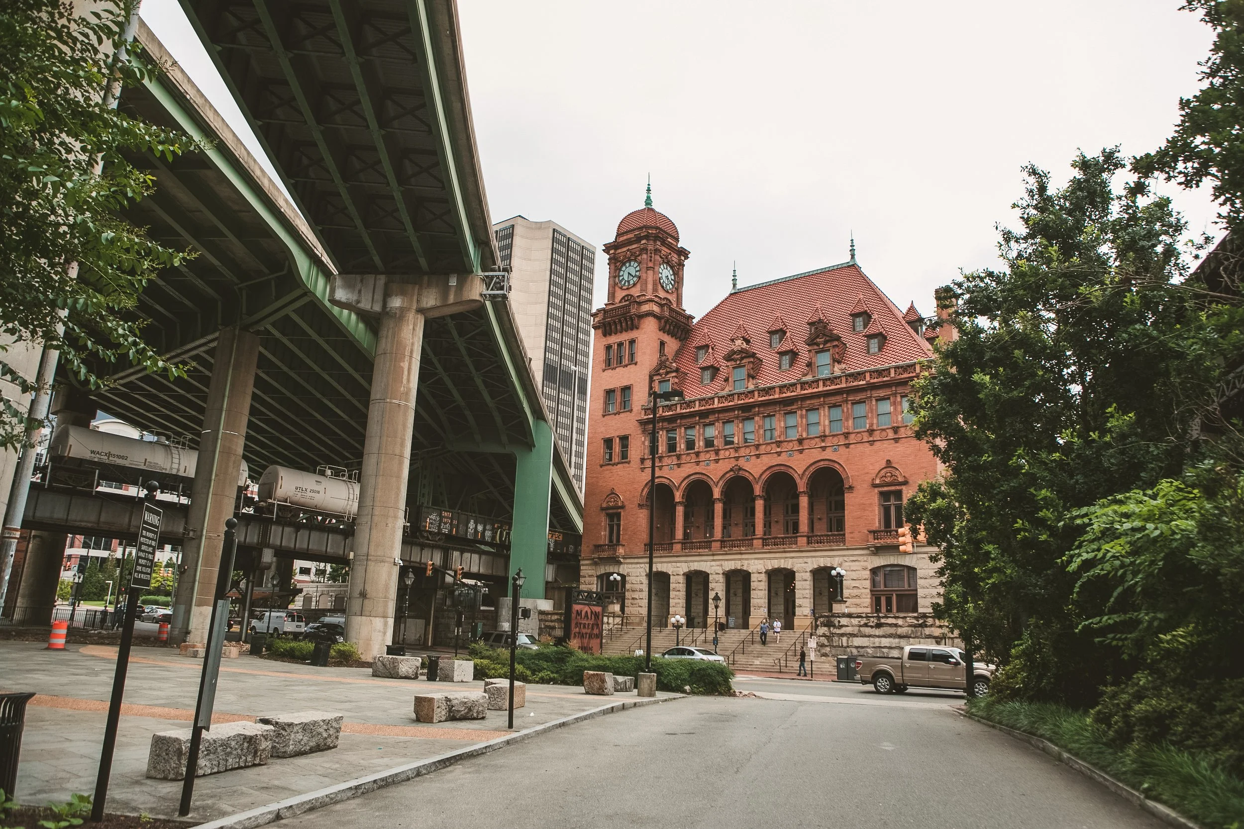 View of an urban area with a historic red brick building featuring a clock tower, an elevated green train track, and modern tall buildings in the background. Trees and street elements also visible.
