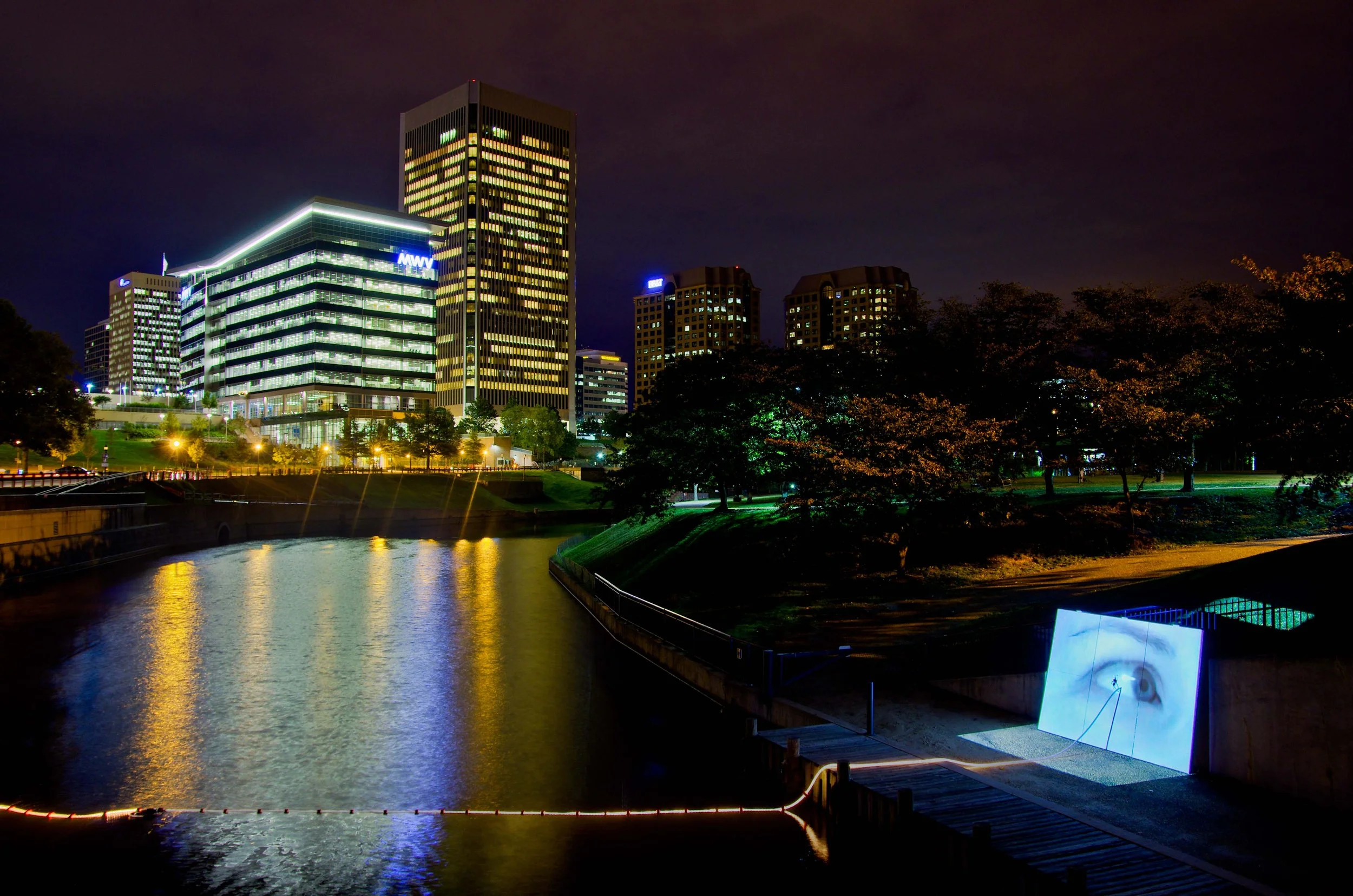 Night view of illuminated modern office buildings along a river with trees and a park in the foreground, and a digital display board on the right side, reflecting some lights on the water.