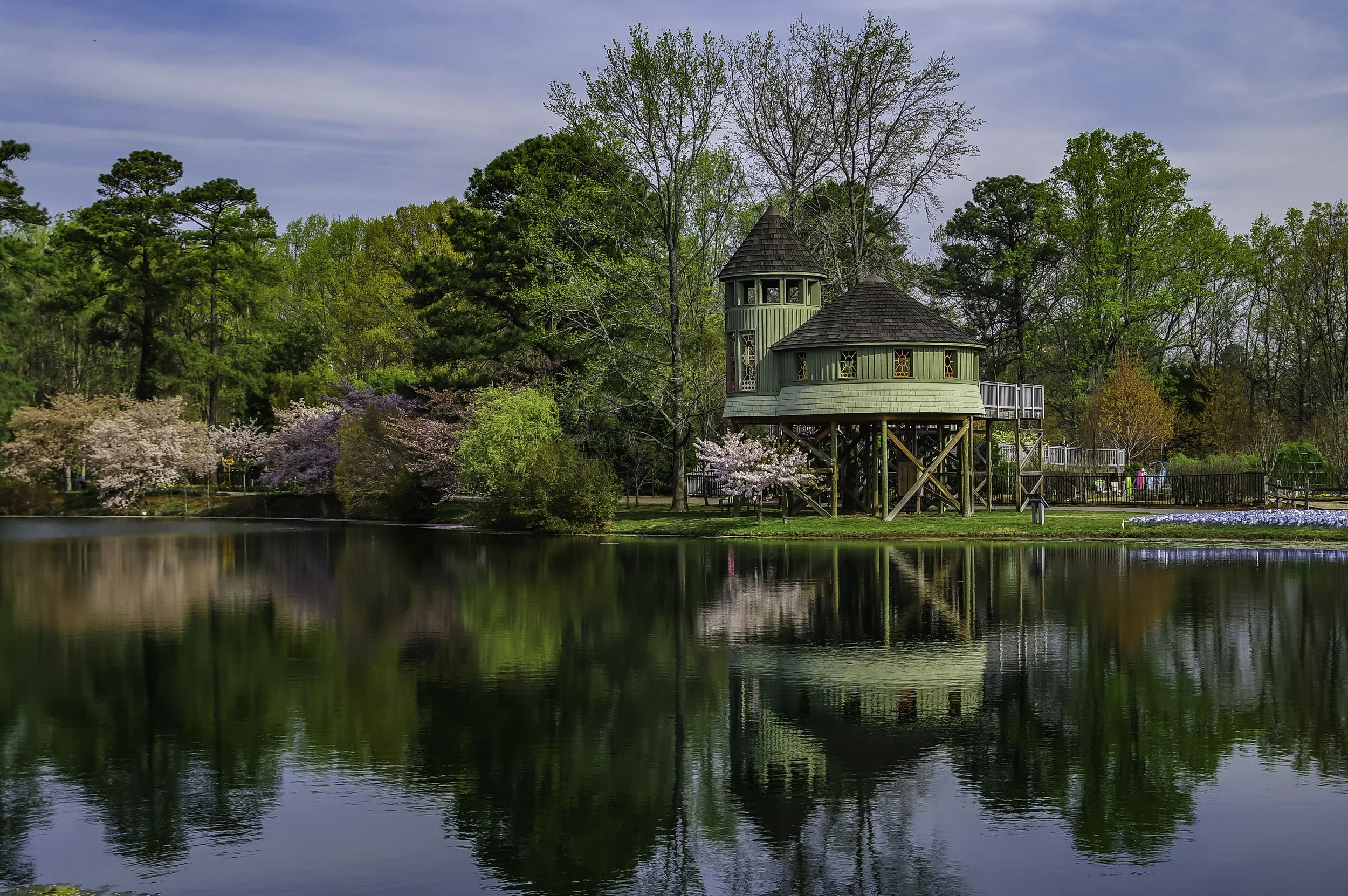 A small green wooden house with a round tower and pointed roof, located by a lake, surrounded by trees and blossoming cherry trees, under a partly cloudy sky.