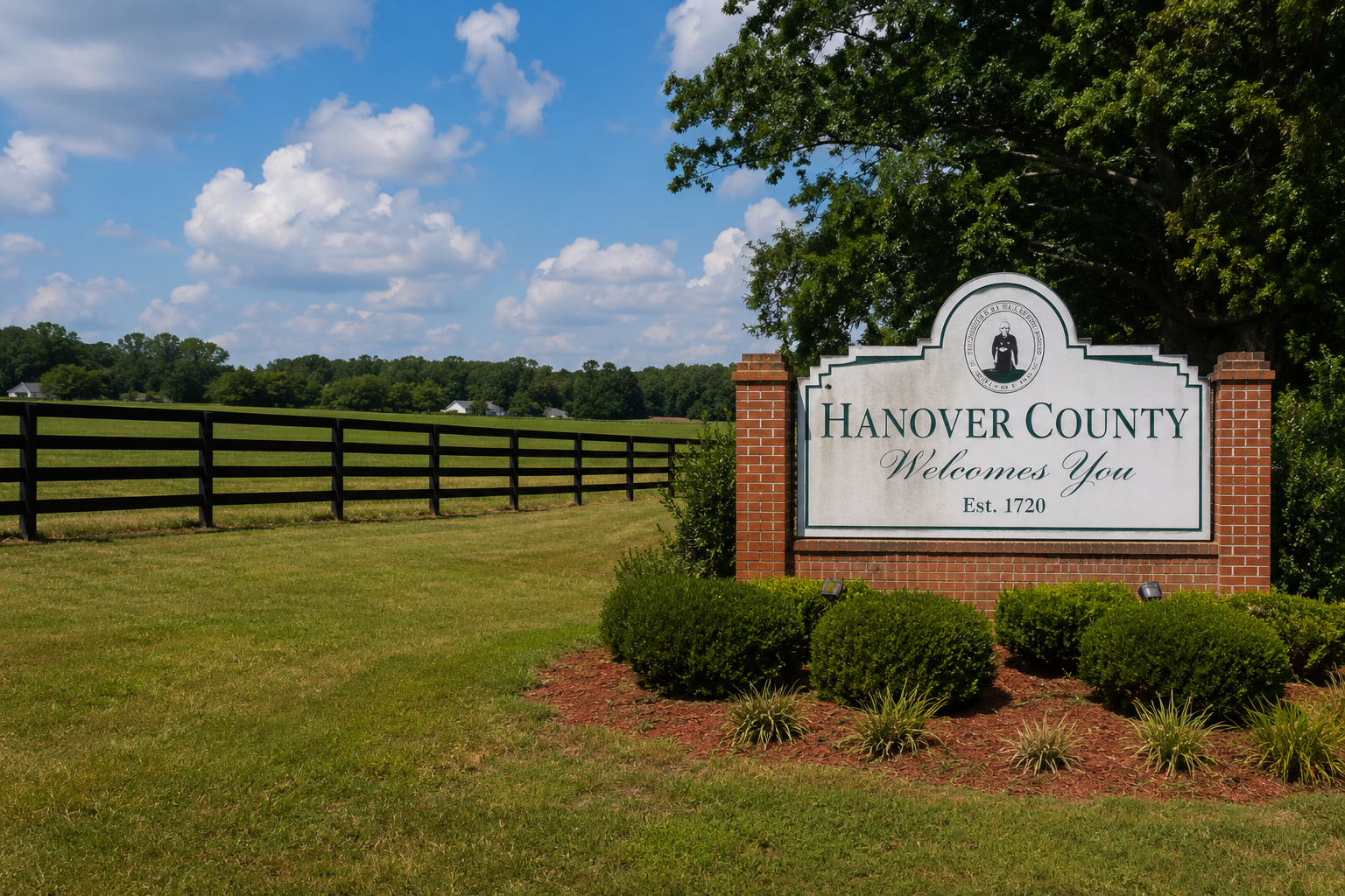A white sign with green and black writing that reads 'Hanover County Welcomes You Est. 1720' next to a brick border and surrounded by green bushes and grass. In the background, there is a black wooden fence, green trees, and a blue sky with white clouds.