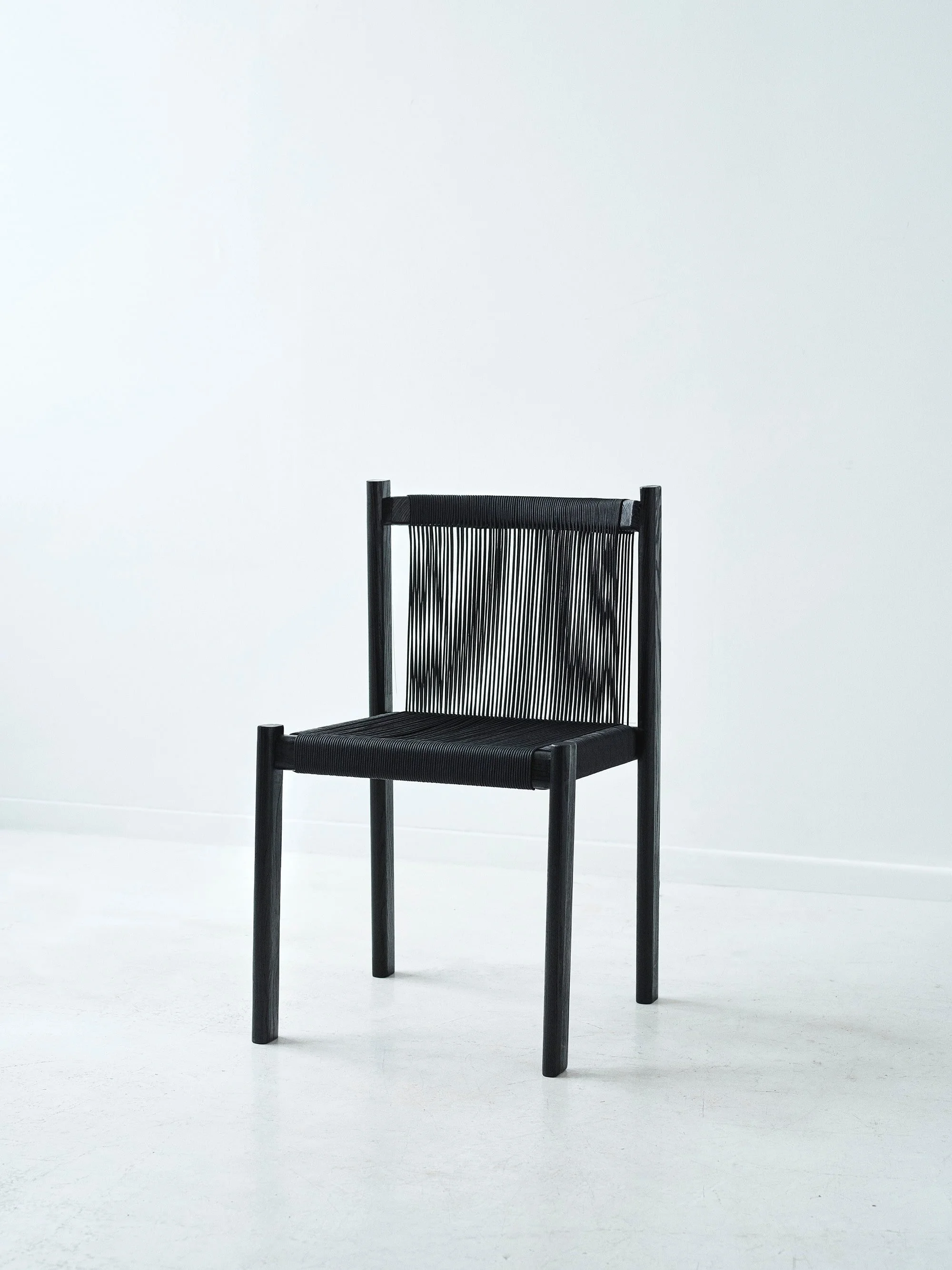 A black modern chair with vertical string backrest and four legs, placed against a plain white wall and white floor.
