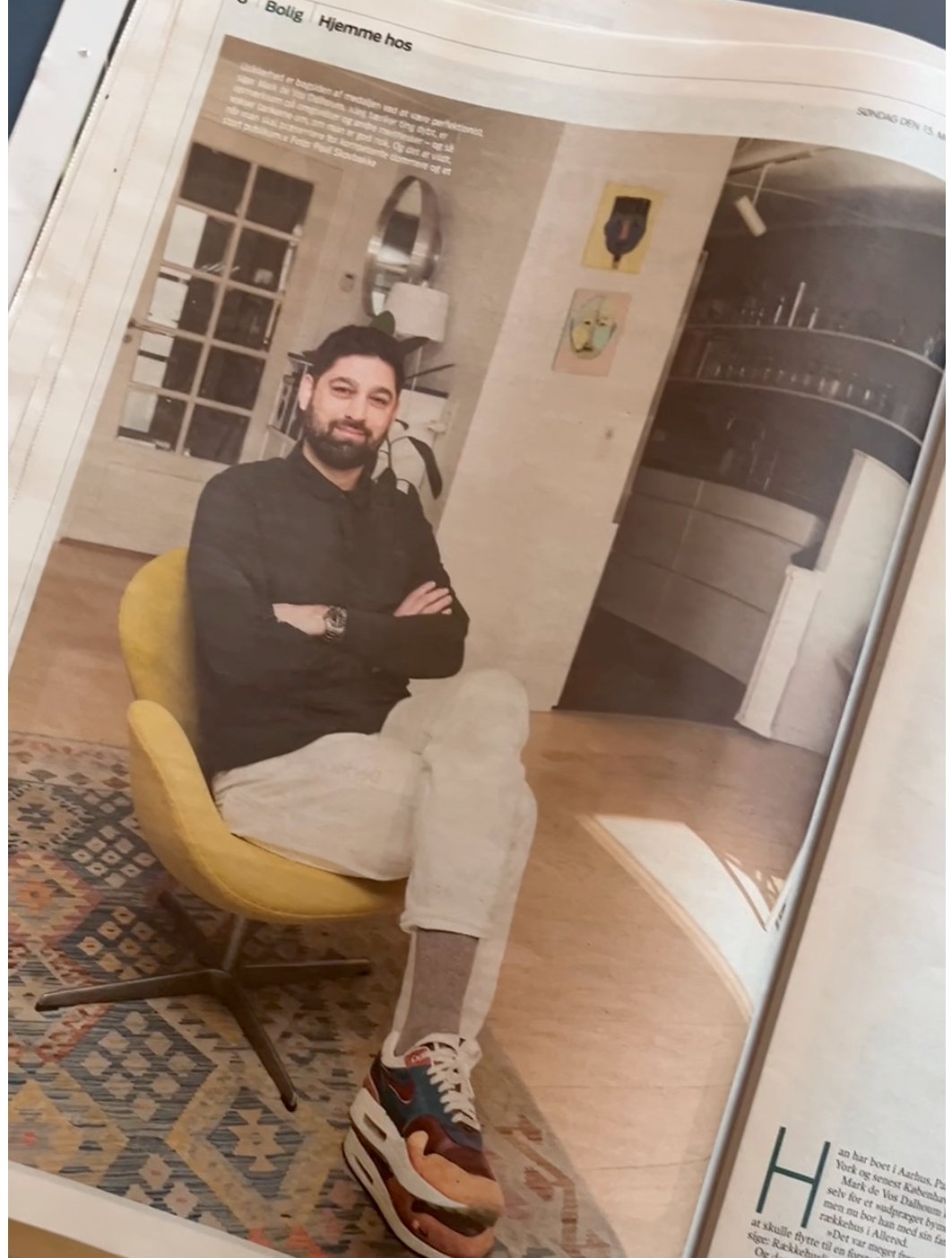 A man with a beard sitting on a yellow chair with crossed arms in a modern, well-lit room. He is wearing a black shirt, beige pants, and sneakers and is smiling at the camera. The room features art on the wall, a window, and a carpeted floor.