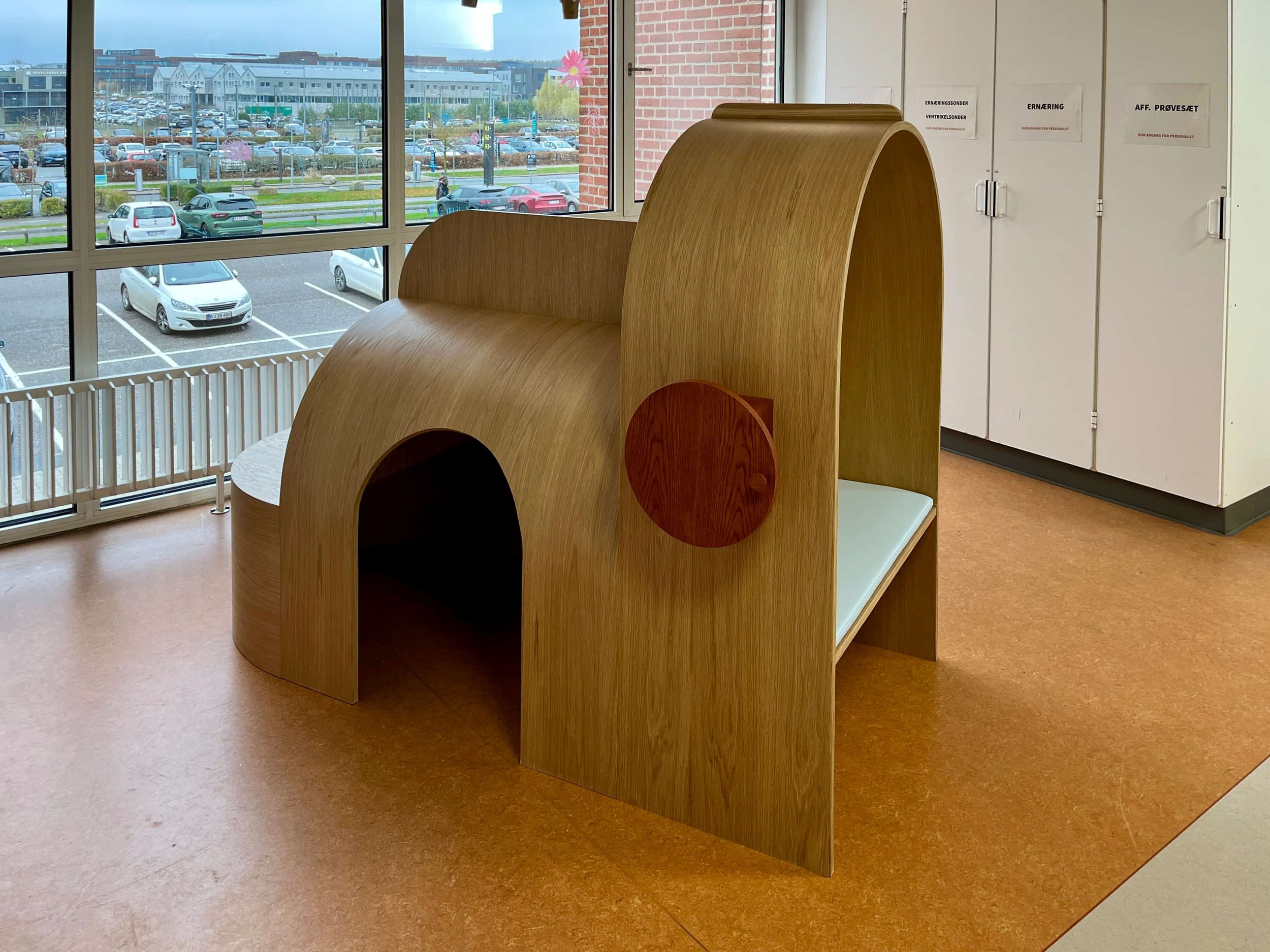 A wooden pet house with a round entrance, located indoors near a window with a parking lot view.