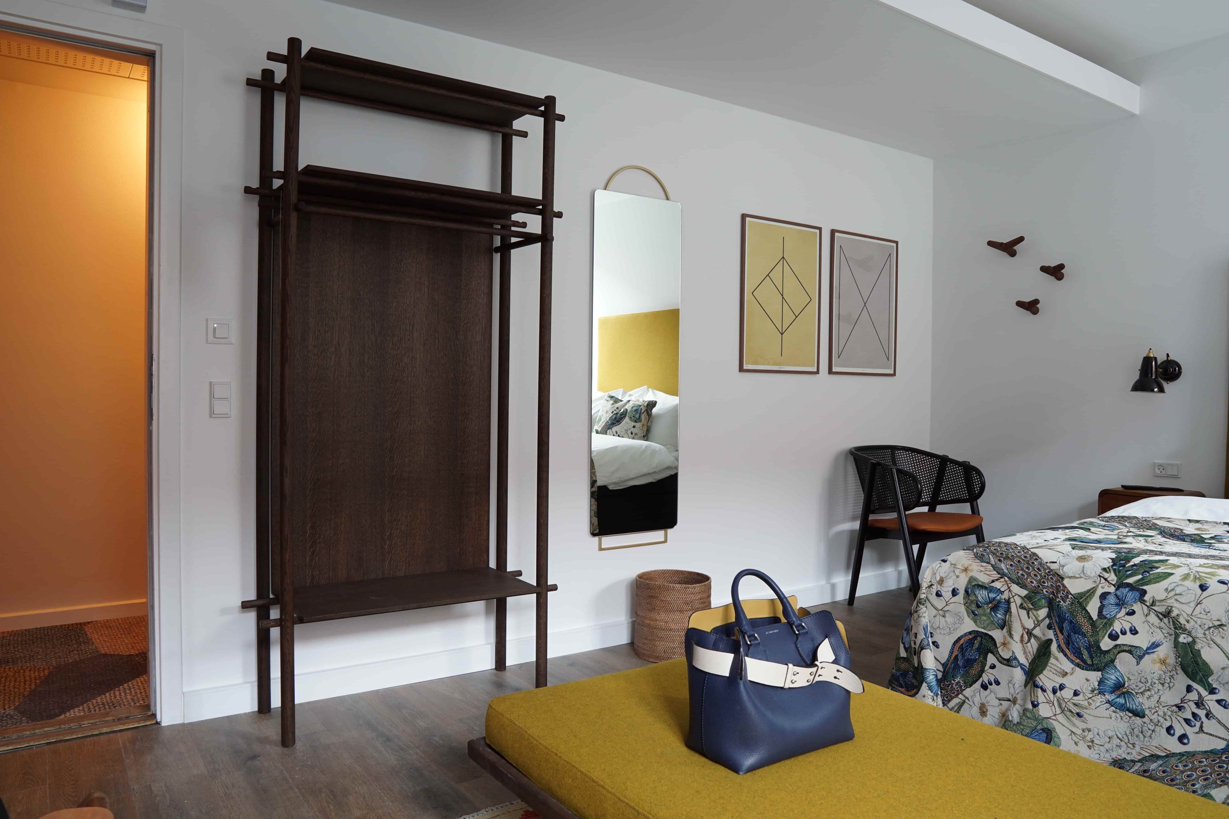 A room with white walls, featuring a bed with floral bedding, two framed abstract artworks, a mirror, and furniture including a dark wooden shelf, black chairs, and a blue handbag on a yellow ottoman.