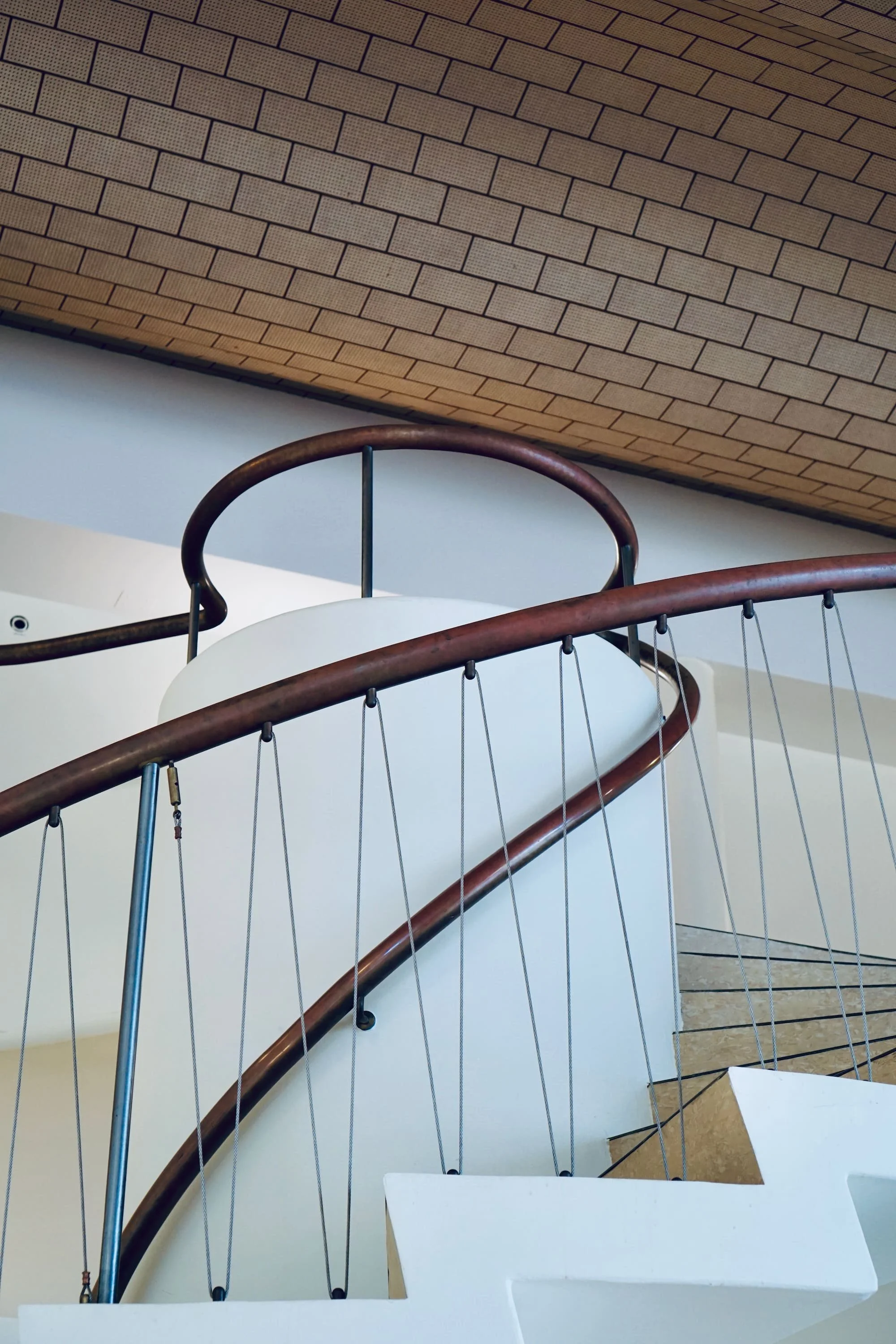 Photo of a modern staircase with white steps, curved wooden handrails, metal supports, and a ceiling with a brick pattern.