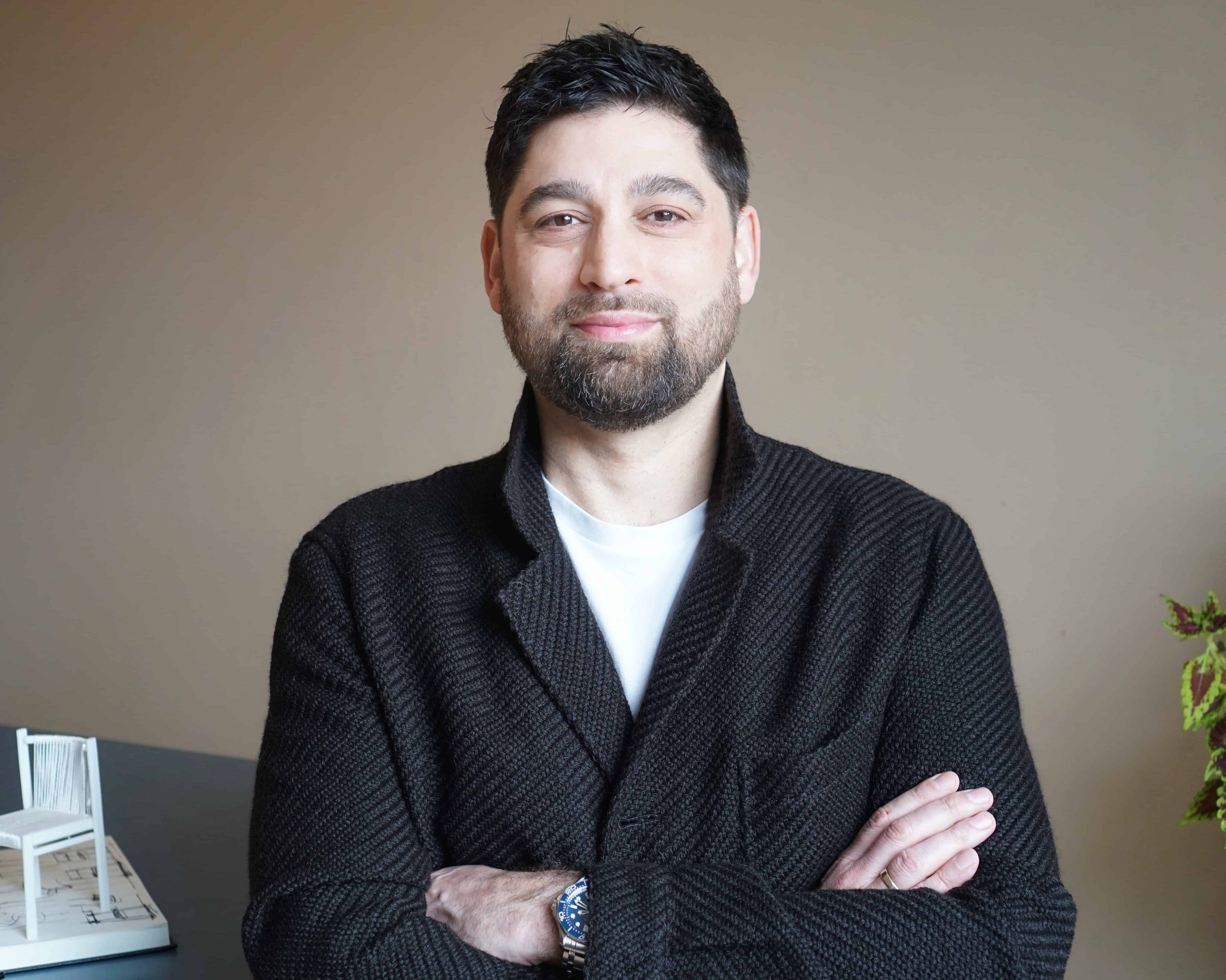 Mark de Vos Dalhoum. A man with dark hair and a beard, wearing a black sweater and blue watch, standing with arms crossed, smiling slightly against a beige background with a white chair and a small red plant nearby.