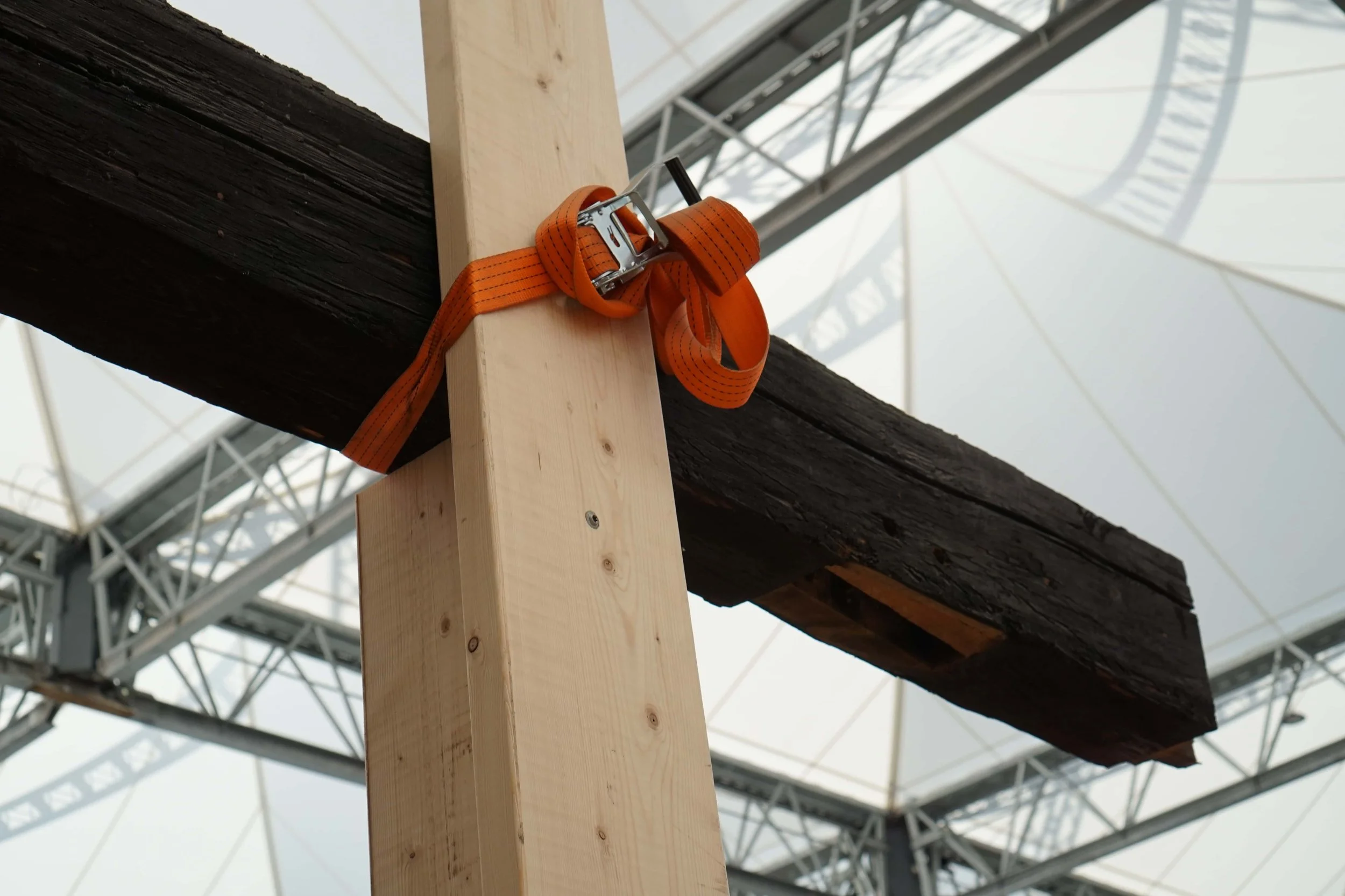 Close-up of wooden construction with an orange ratchet strap securing a black beam in place, with metal scaffolding or structural framework in the background. Børsen, Copenhagen after the fire.