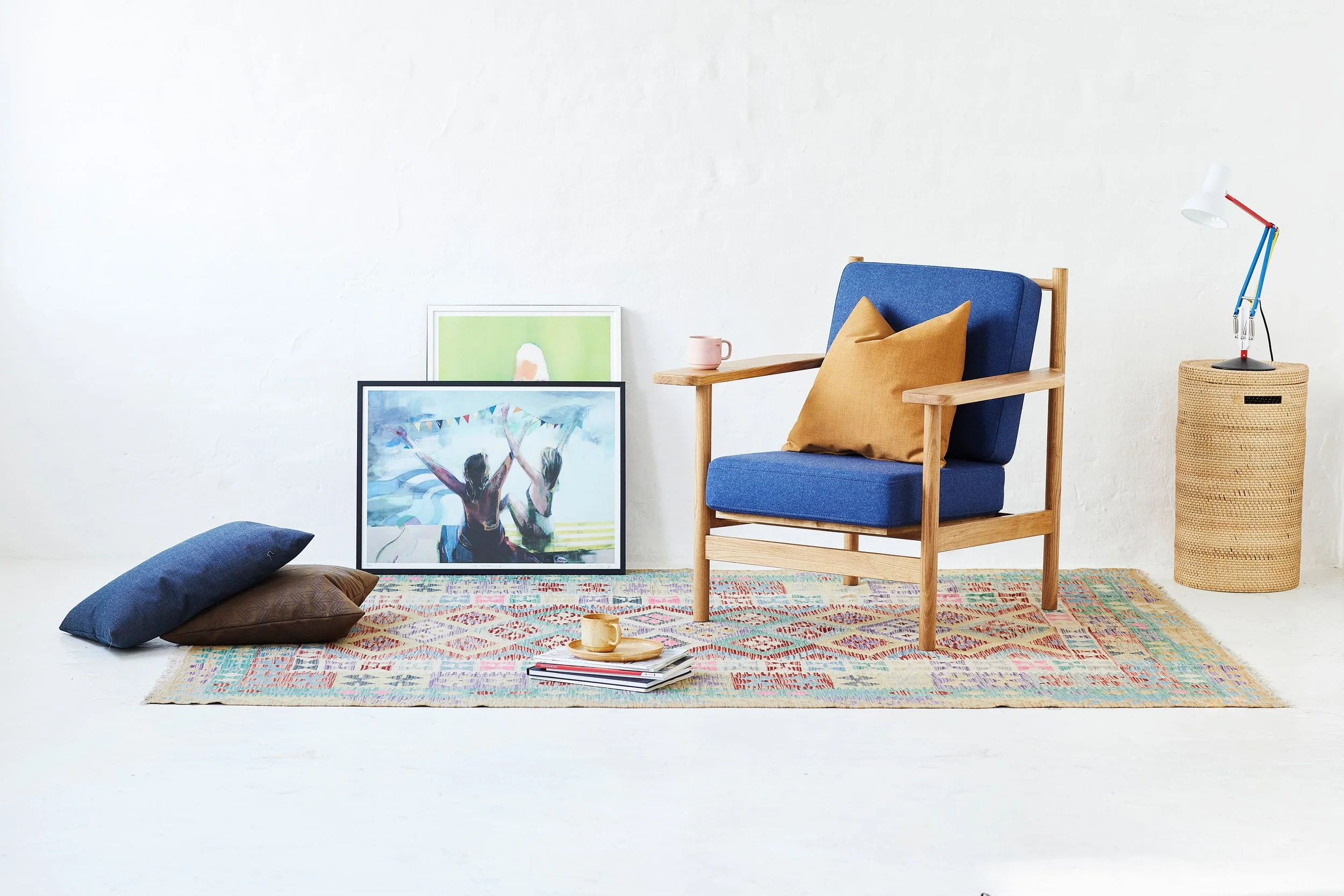 A cozy interior scene with a blue armchair with a tan pillow, a pink mug on the armrest, and framed artwork leaning against a plain white wall. There's a woven basket with a colorful adjustable lamp on it, and a patterned rug on the floor with two pillows and a neatly stacked set of magazines and a cup.