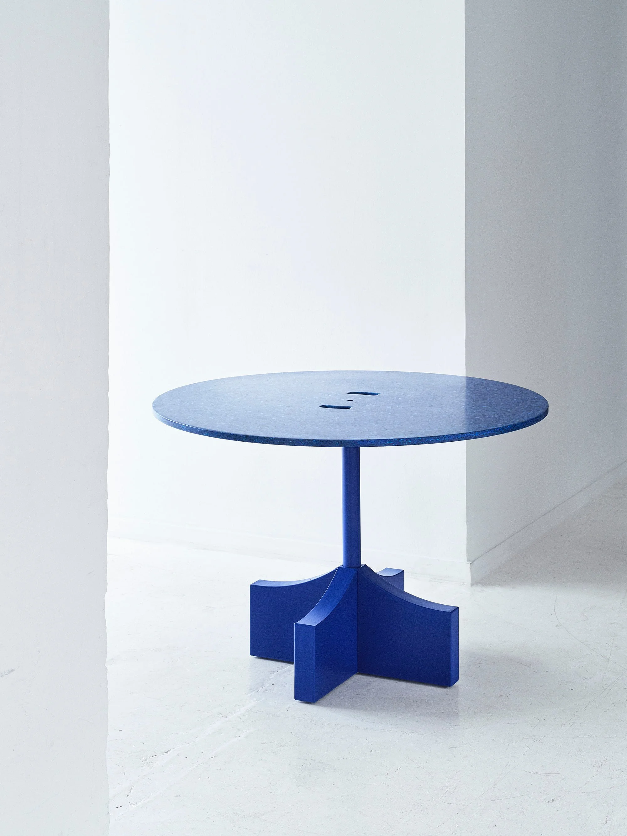 A blue table with a rounded top and a single central leg, standing on a white floor against white and grey walls.