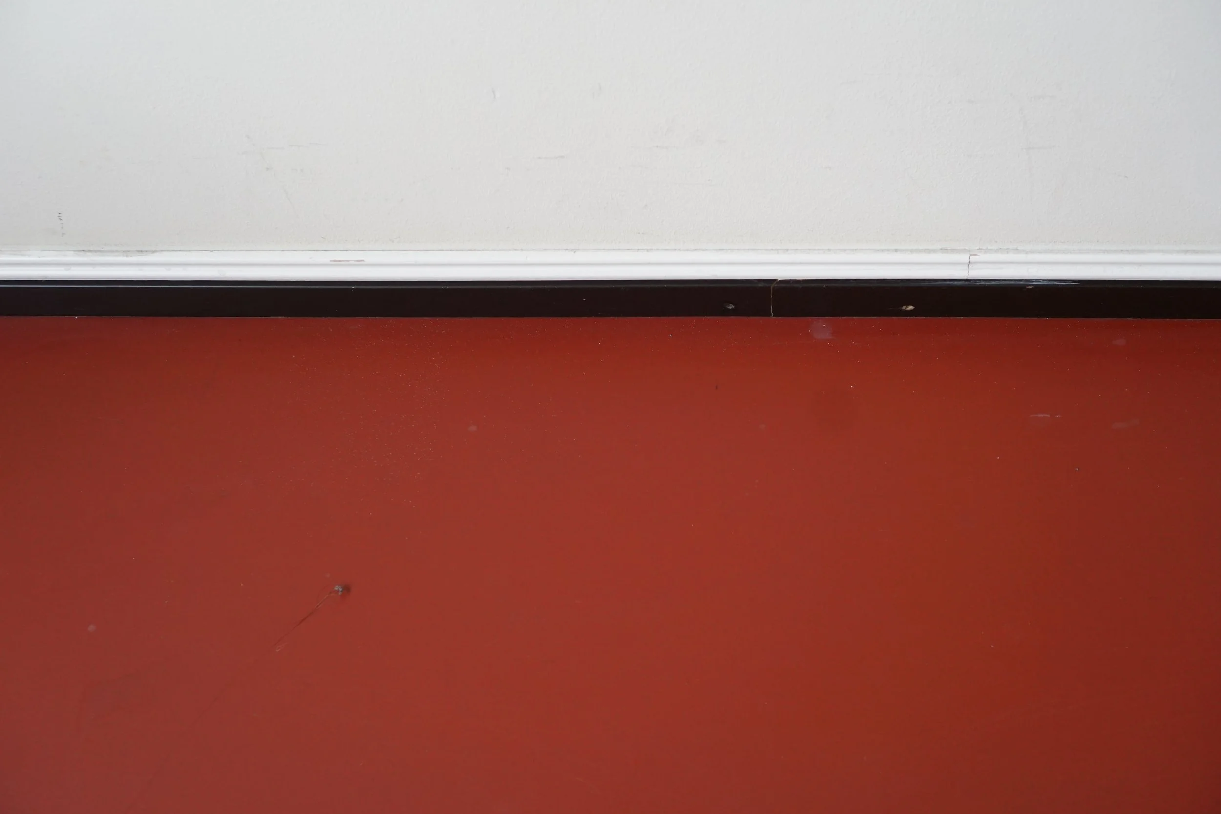 View of a baseboard heater on a white wall above a red floor, with a small crack in the wall near the baseboard.