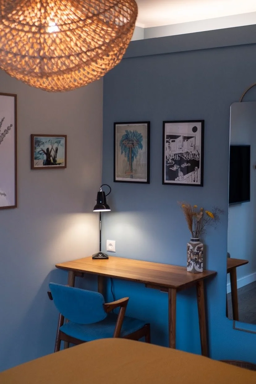 Living room corner with a wooden desk, blue chair, black desk lamp, wall art, a vase with dried flowers, and a woven ceiling light.