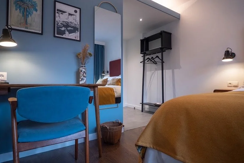 A cozy hotel room featuring a bed with mustard-colored bedding, a mirror, a black clothing rack, and a small desk with a blue chair. Decor includes framed art and a vase with dried plants.