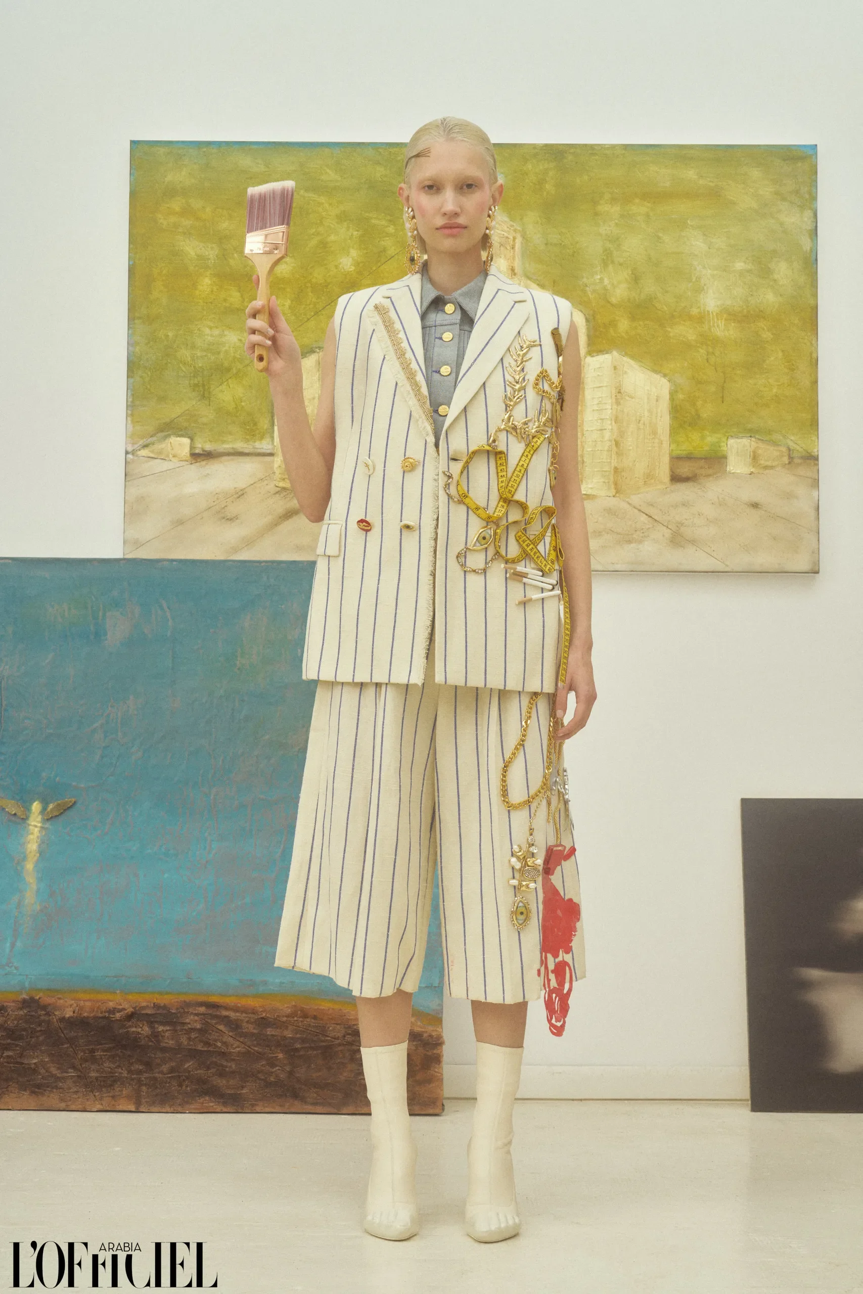 Modella in studio d'arte con completo gessato composto da gilet e bermuda decorati da metri da sarta e catene, con pennello da pittura in mano.