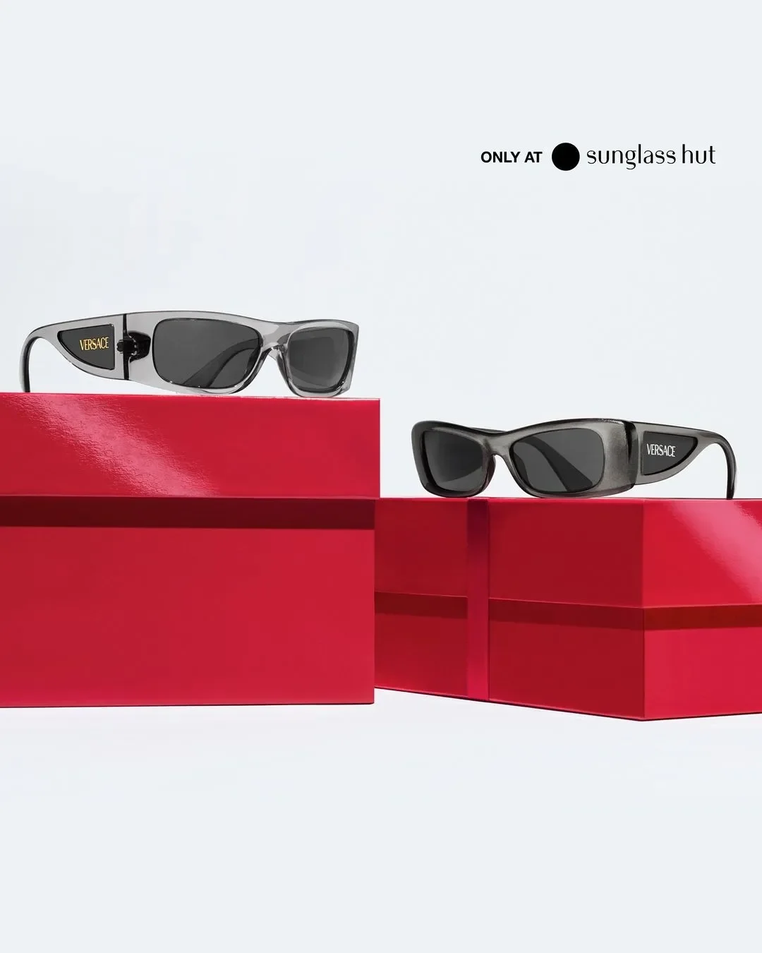 Fotografia still life per Sunglass Hut con due paia di occhiali da sole rettangolari Versace in acetato grigio trasparente con logo sulle aste, disposti su grandi pacchi regalo rossi natalizi.