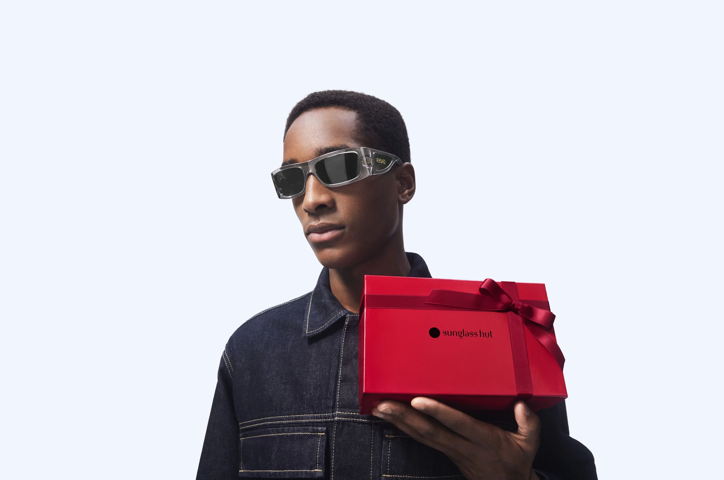 Modello maschile in giacca di jeans che indossa occhiali da sole rettangolari Versace in acetato grigio trasparente, mentre tiene in mano una scatola regalo rossa brandizzata Sunglass Hut.