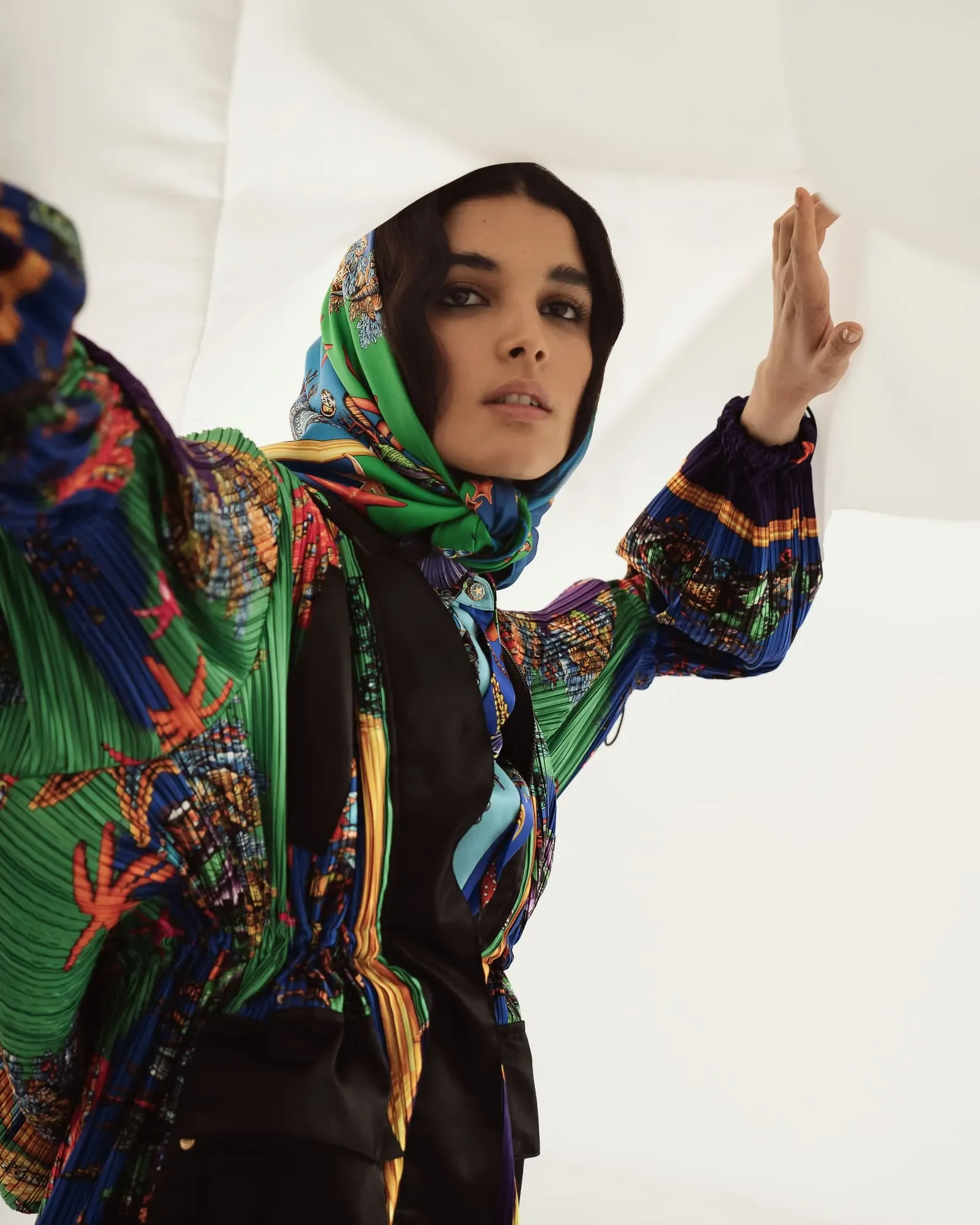 Primo piano espressivo della modella con foulard e capospalla a motivi stampati multicolor mentre solleva un velo scenografico.