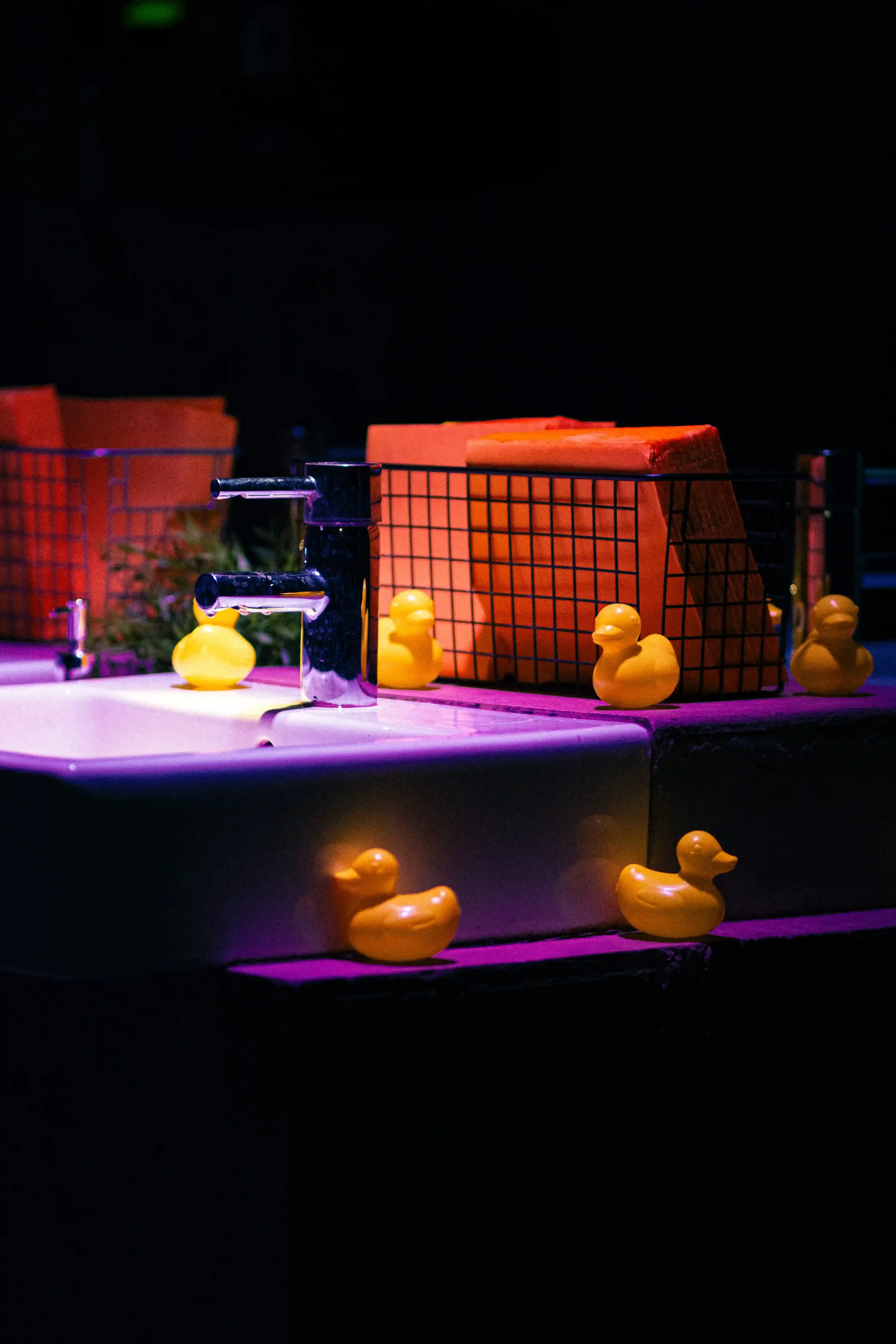 Dettaglio di set design d'atmosfera per l'area bagno dell'evento Mandarina Duck a Berlino, con paperelle di gomma gialle allestite sui lavabi e luci al neon viola.
