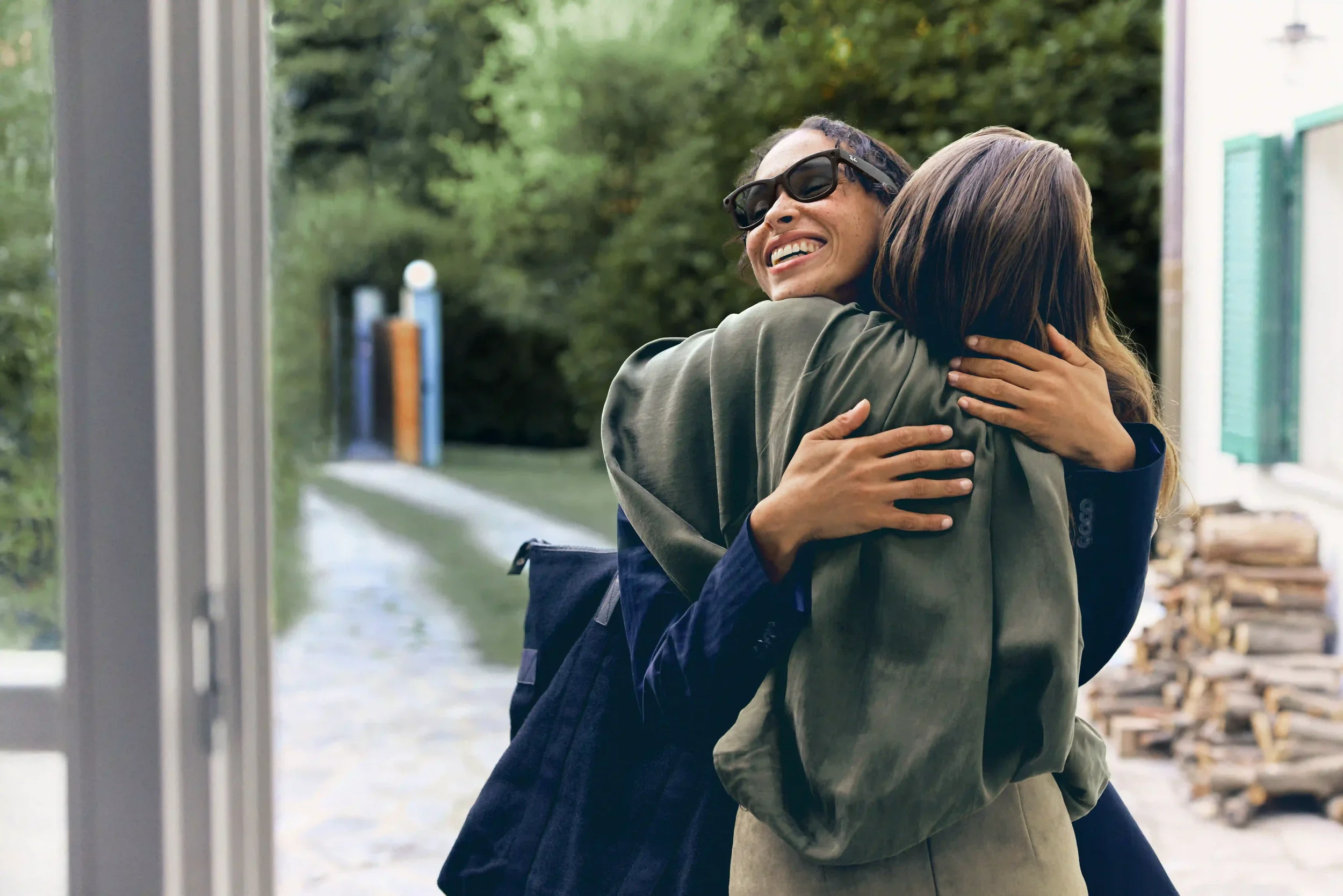 Scatto emozionale per la campagna di Natale Sunglass Hut con due amiche che si abbracciano calorosamente all'esterno della casa, con modella che indossa occhiali da sole neri squadrati.