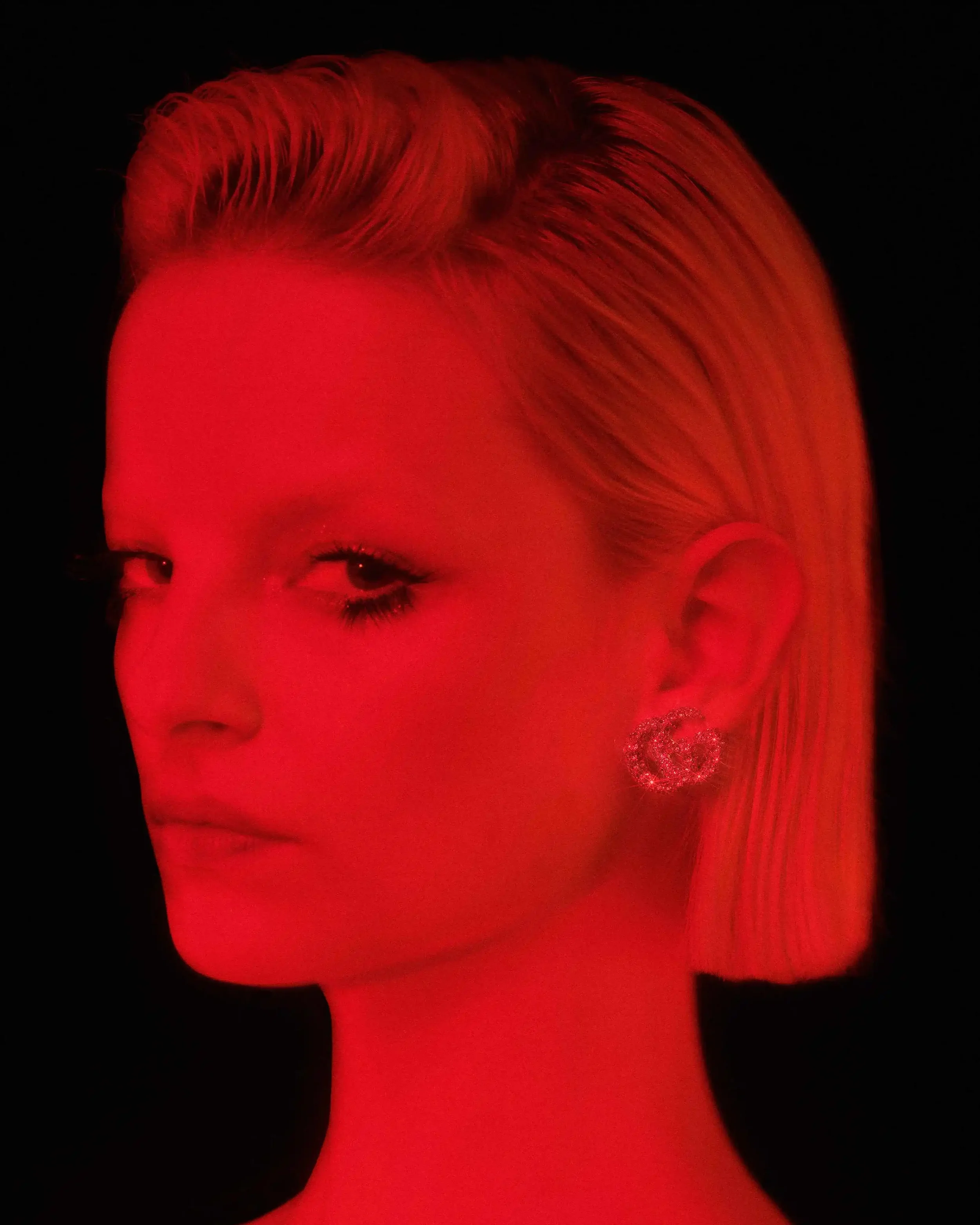Primo piano beauty in luce rossa intensa per DNA Magazine e Gucci. Modella con taglio corto biondo e make-up occhi marcato, con in evidenza l'orecchino a lobo con il logo GG Gucci tempestato di cristalli.