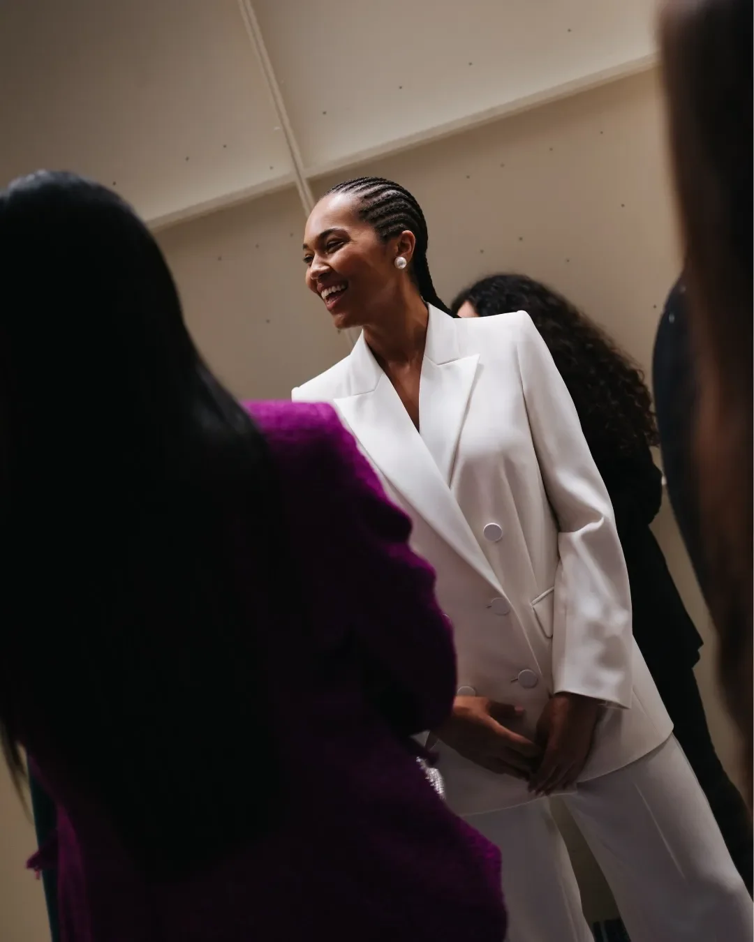 Scatto spontaneo nel backstage della sfilata Marina Rinaldi: modella sorridente con acconciatura a treccine indossa un tailleur pantalone doppio petto bianco con bottoni decorativi.