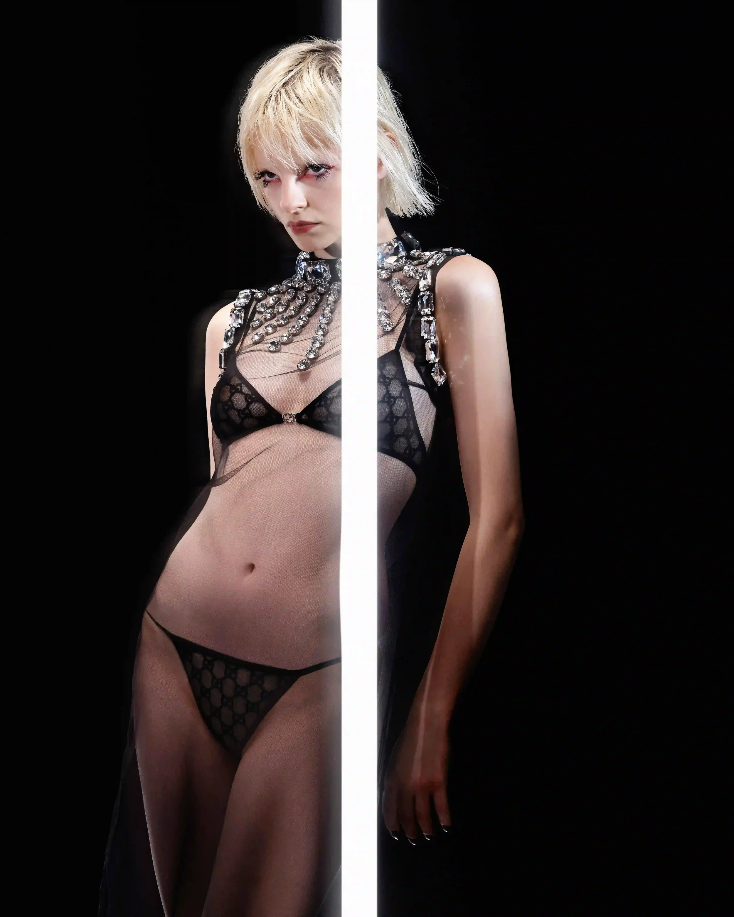 Ritratto editoriale ad alto impatto visivo per DNA Magazine in collaborazione con Gucci. Modella bionda in posa divisa da una barra di luce neon bianca verticale su sfondo nero, con indosso un completo intimo nero con monogramma GG Gucci, abito scivo