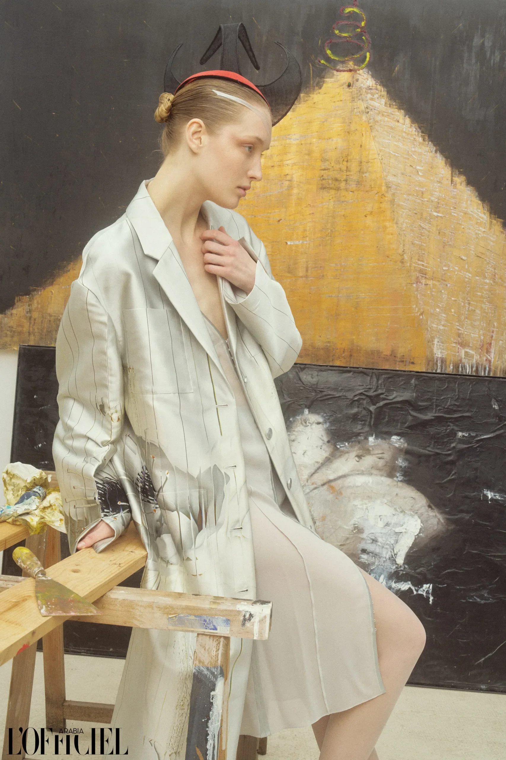 Modella seduta in uno atelier d'arte accanto a un cavalletto di legno, con blazer oversize grigio gessato e originale cappello scultoreo nero e rosso.