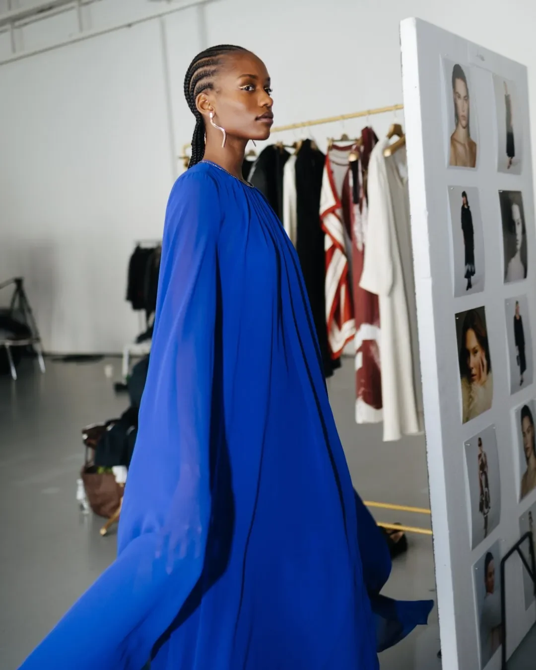 Modella nel backstage in un abito lungo e fluido blu elettrico stile mantella, mentre sfila accanto allo stender della collezione e alla parete dei bozzetti e casting Marina Rinaldi