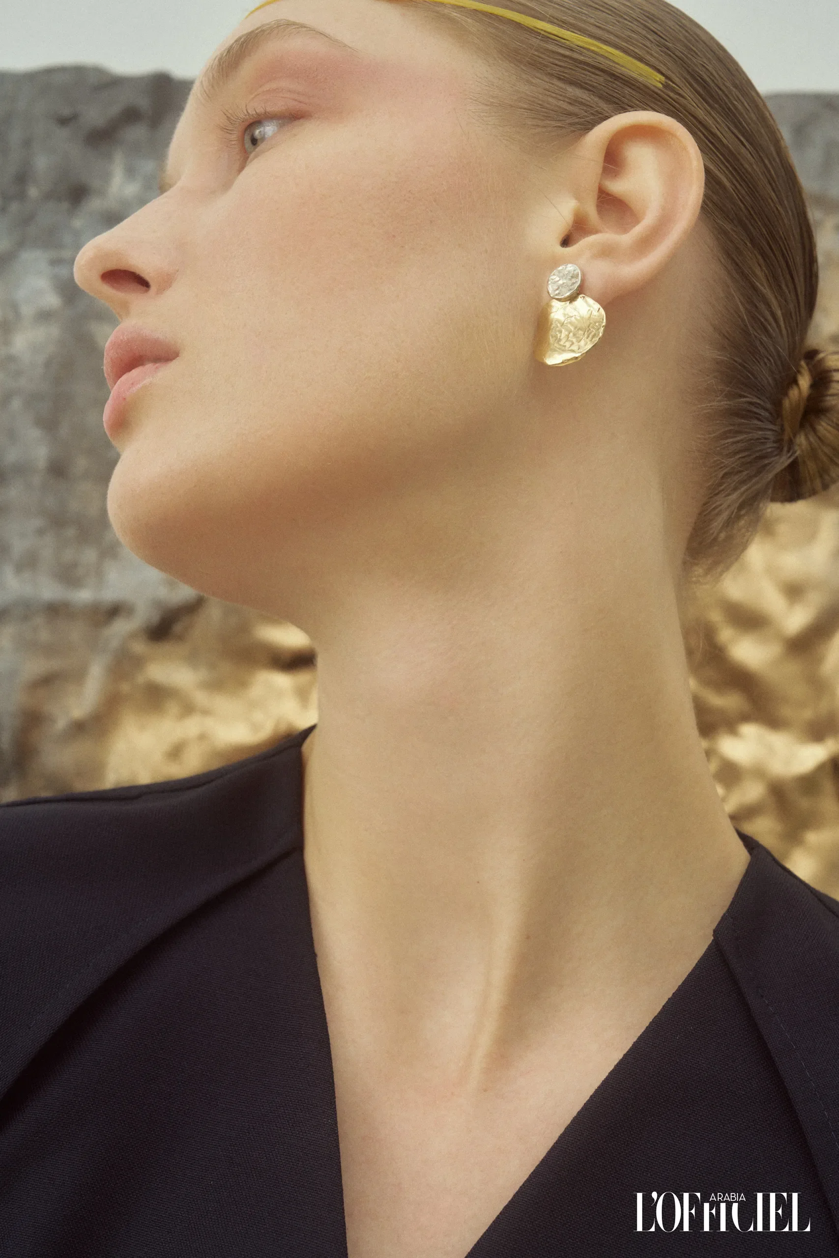 Dettaglio e primo piano del profilo della modella con orecchino scultoreo in oro martellato e abito nero con scollo geometrico.