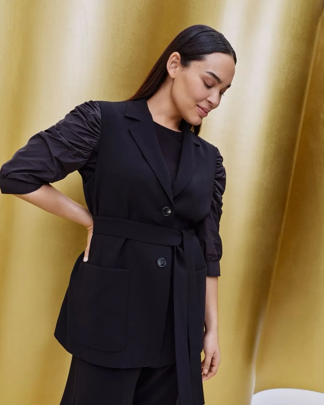 Modella in completo sartoriale nero con cintura coordinata e maniche arricciate, ritratta di profilo davanti allo sfondo metallico oro del set della sfilata Marina Rinaldi