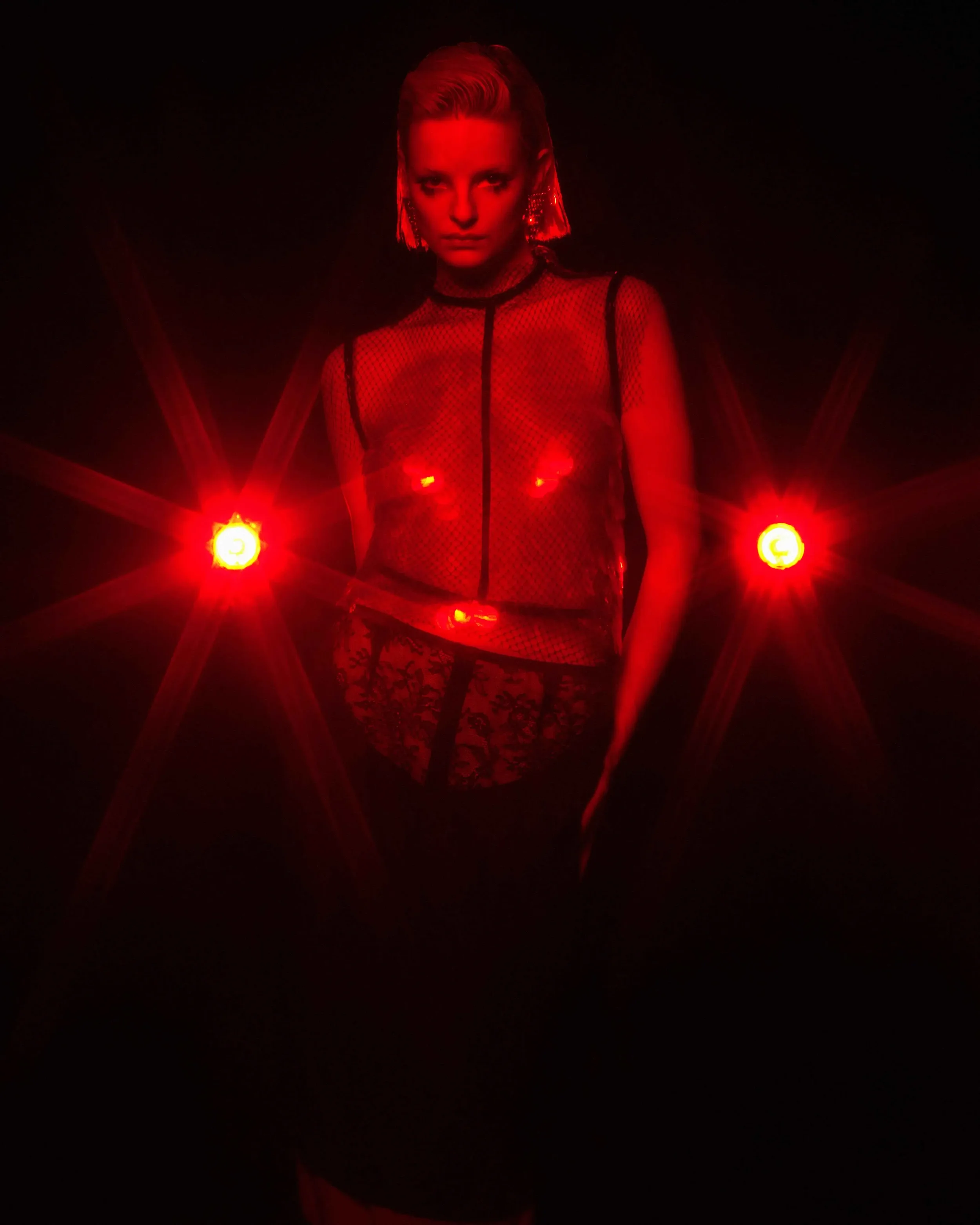 Fotografia editoriale per DNA Magazine in collaborazione con Gucci. Modella in posa al centro della composizione affiancata da due potenti punti luce rossi a stella, con indosso un top semitrasparente in rete nera su intimo in pizzo.