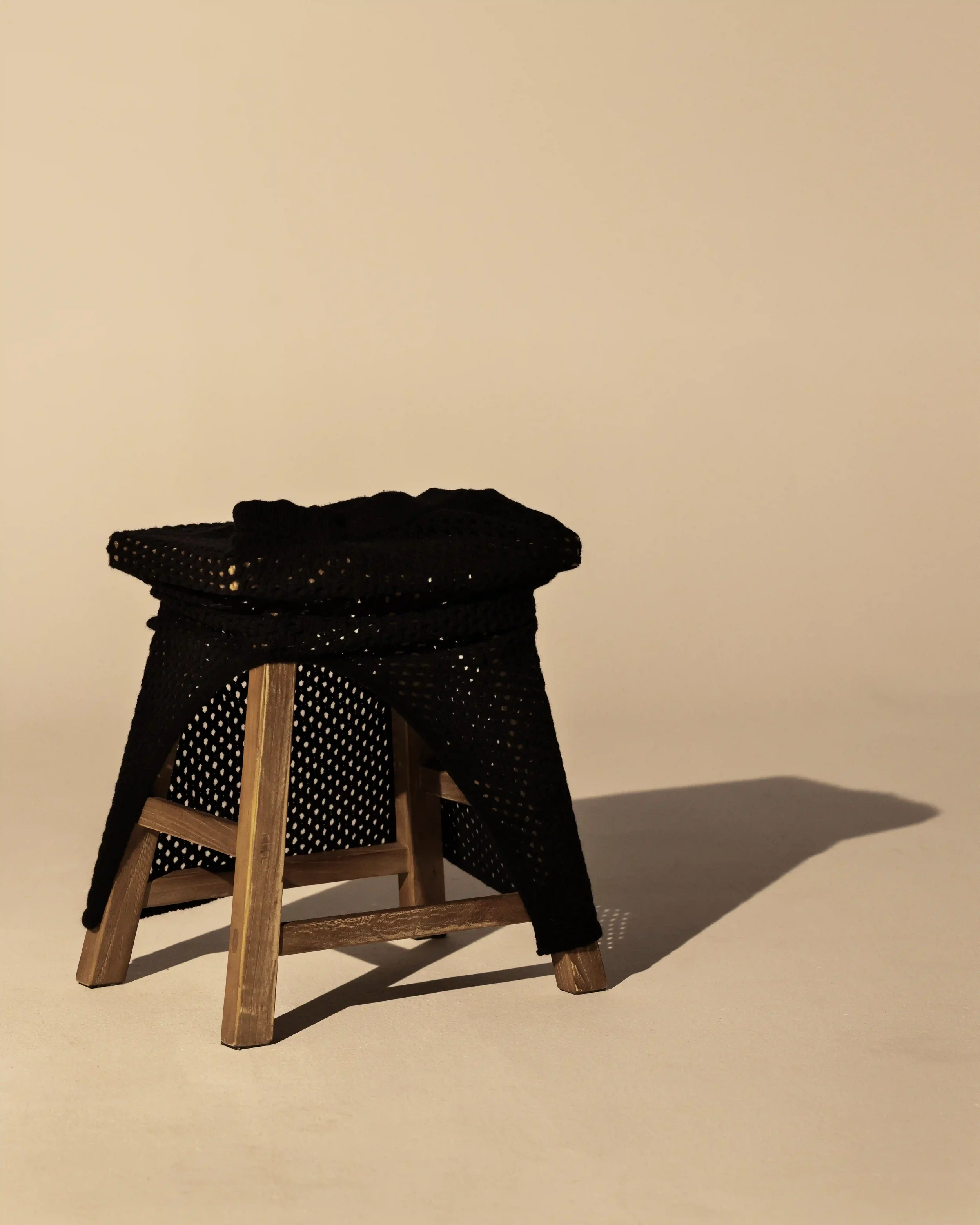 Fotografia still life editoriale per Laneus: maglia traforata nera adagiata su uno sgabello di legno grezzo con ombre definite su sfondo di toni neutri caldi.