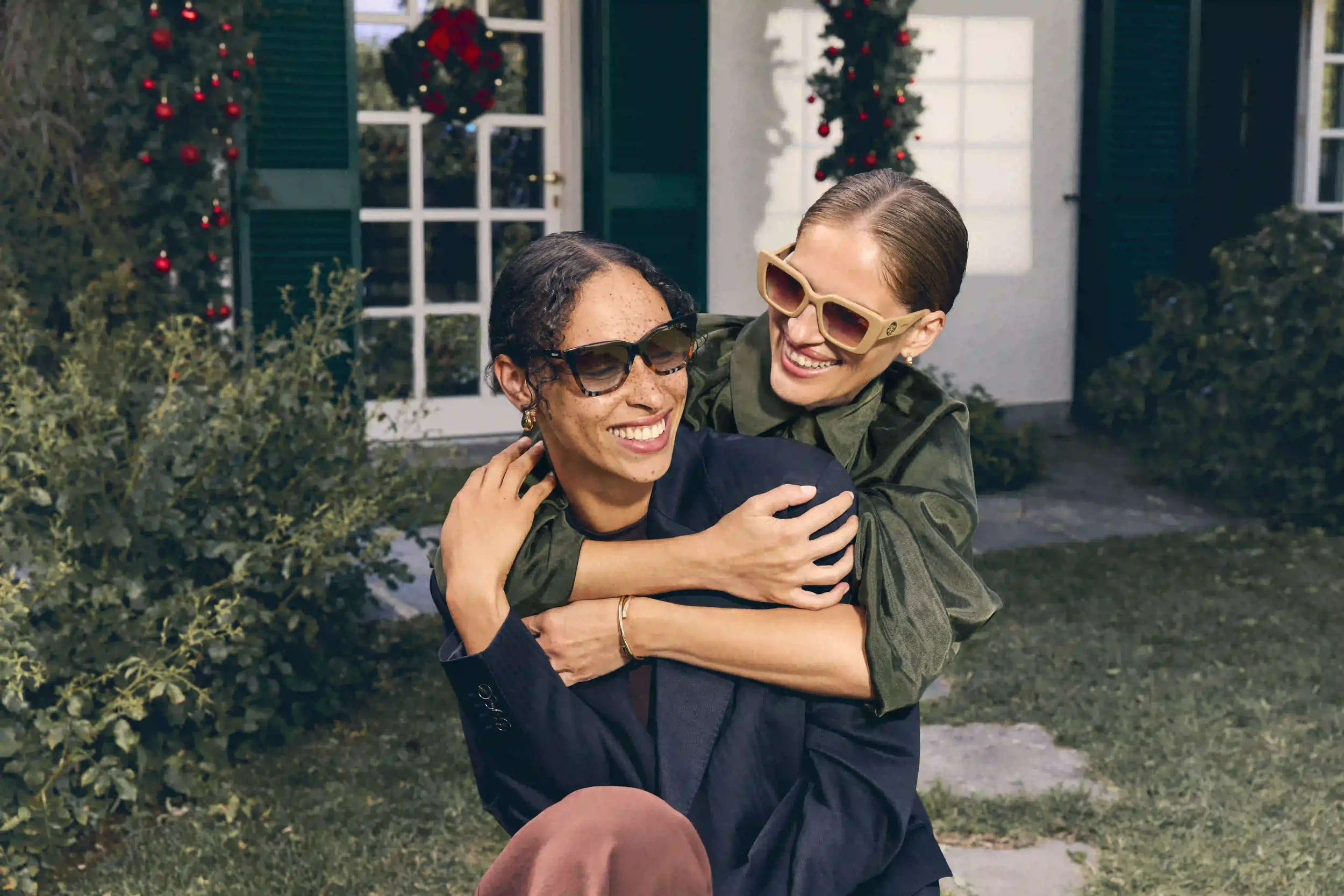 Fotografia di moda outdoor per Sunglass Hut con due modella che si abbracciano sorridenti davanti a una porta allestita per Natale, indossando occhiali da sole di design squadrati e tartarugati.