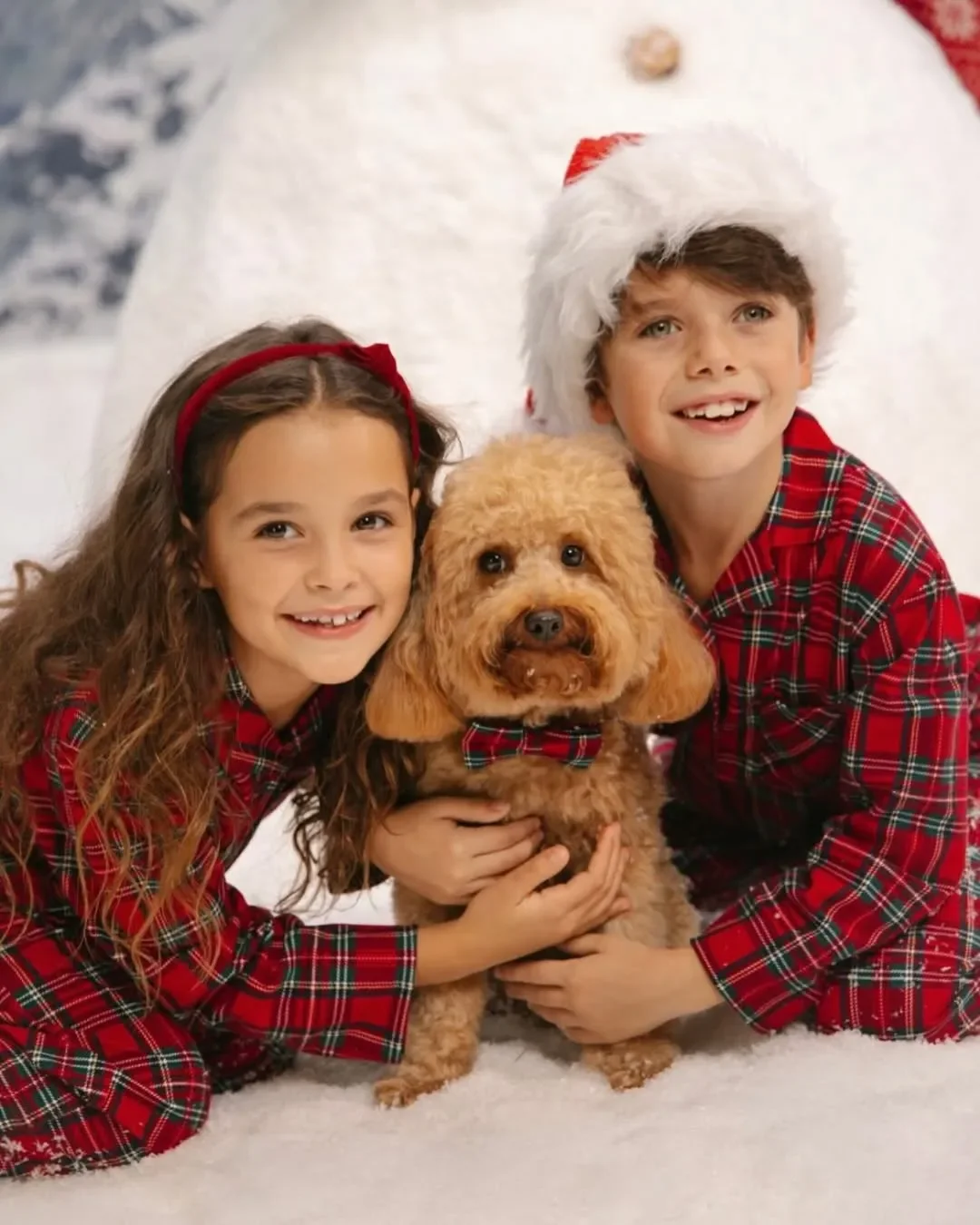 Primo piano di due bambini sorridenti in pigiama natalizio tartan rosso Yamamay che abbracciano un cagnolino con papillon coordinato su un soffice manto di neve artificiale.