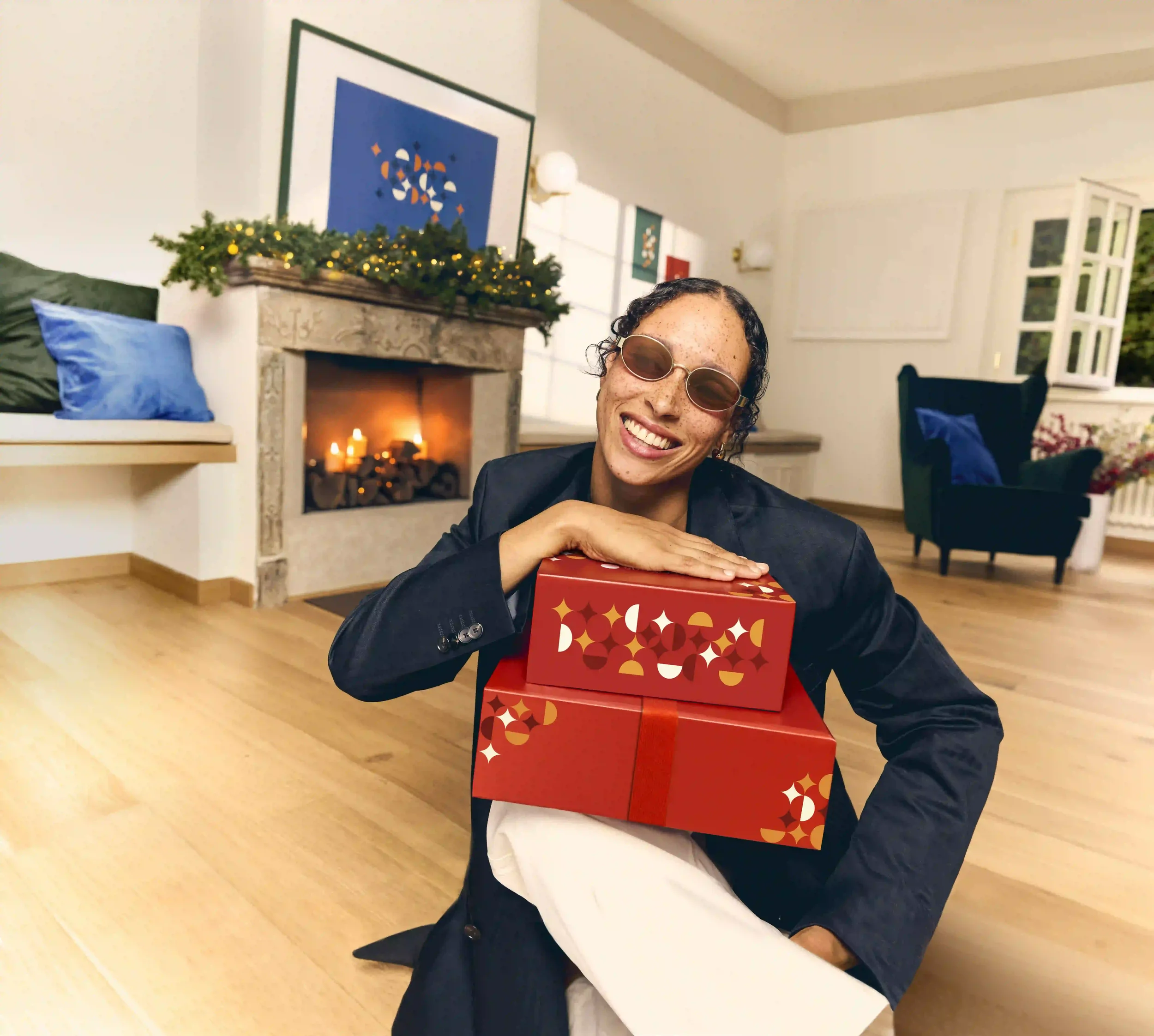 Fotografia editoriale e lifestyle per la campagna natalizia di Sunglass Hut con modella seduta vicino al camino acceso che abbraccia scatole regalo rosse, indossando occhiali da sole ovali con montatura dorata.