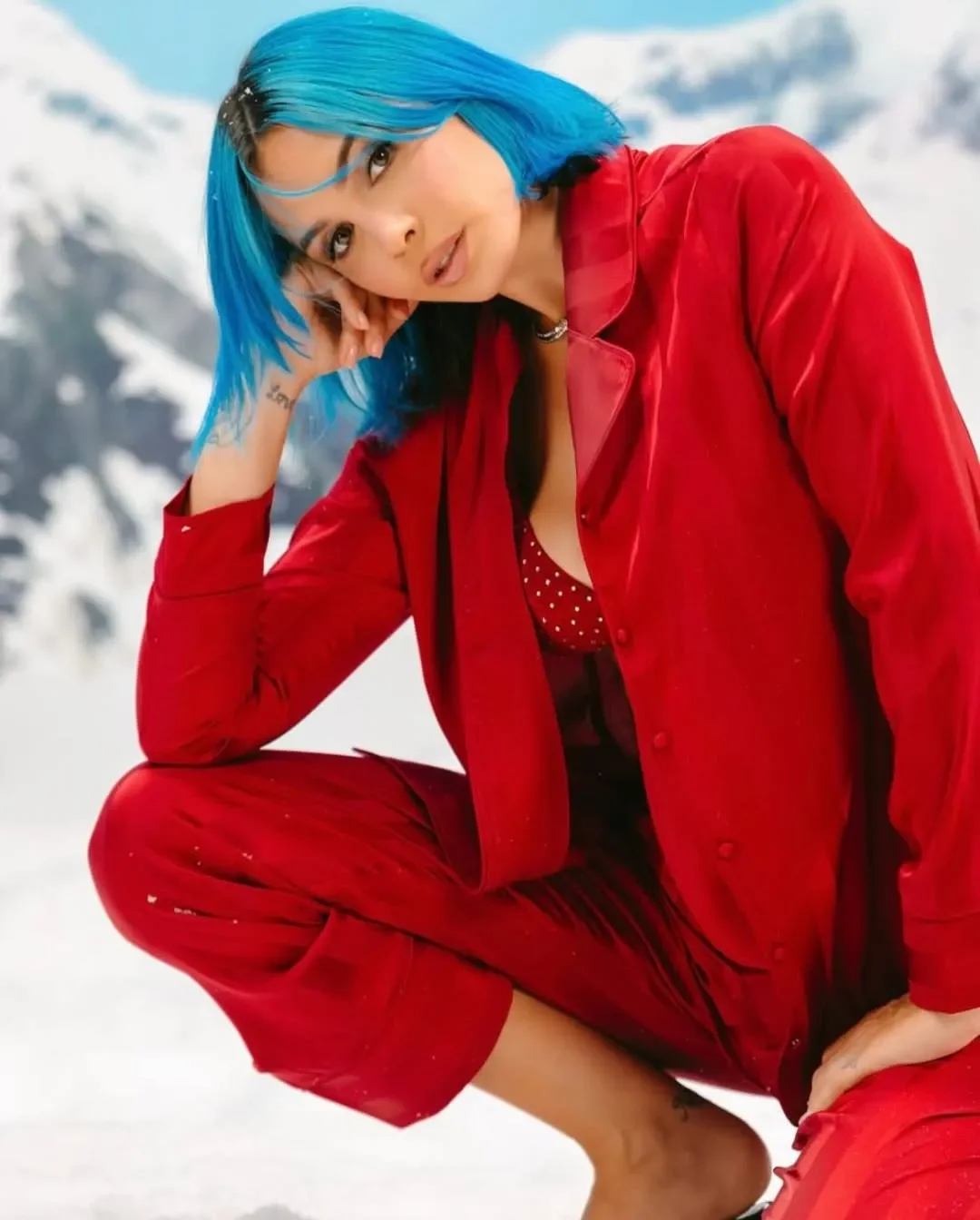 Ritratto in primo piano di Rose Villain accucciata sulla neve con completo pigiama rosso lucido Yamamay e acconciatura blu, in un'atmosfera invernale e natalizia