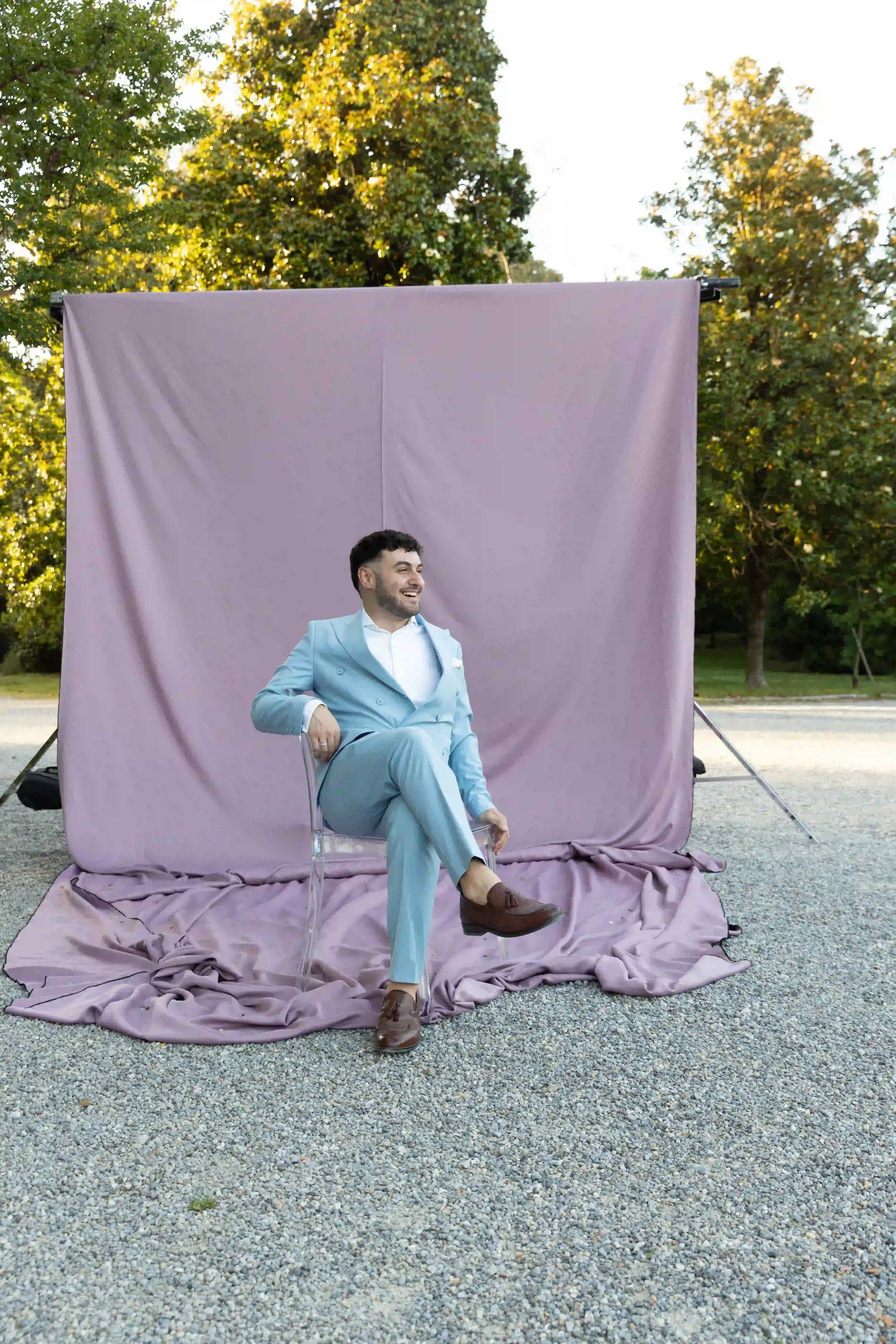 Set fotografico photobooth allestito nel parco della villa con fondale scenografico in tessuto color malva drappeggiato, sedia trasparente Ghost e abito azzurro da cerimonia
