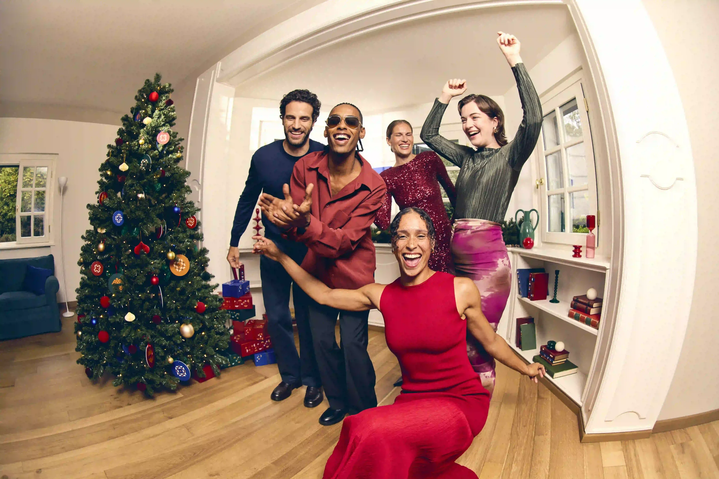 Fotografia di gruppo corale per la campagna natalizia Sunglass Hut con cinque ragazzi che festeggiano e sorridono davanti all'albero di Natale decorato