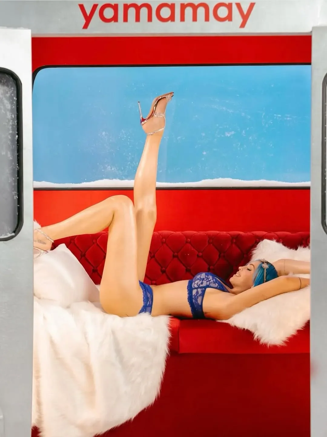 Rose Villain sdraiata su un divano in velluto rosso all'interno della cabinovia Yamamay, indossa un completo intimo in pizzo blu elettrico e scarpe con tacco alto per la campagna di Natale