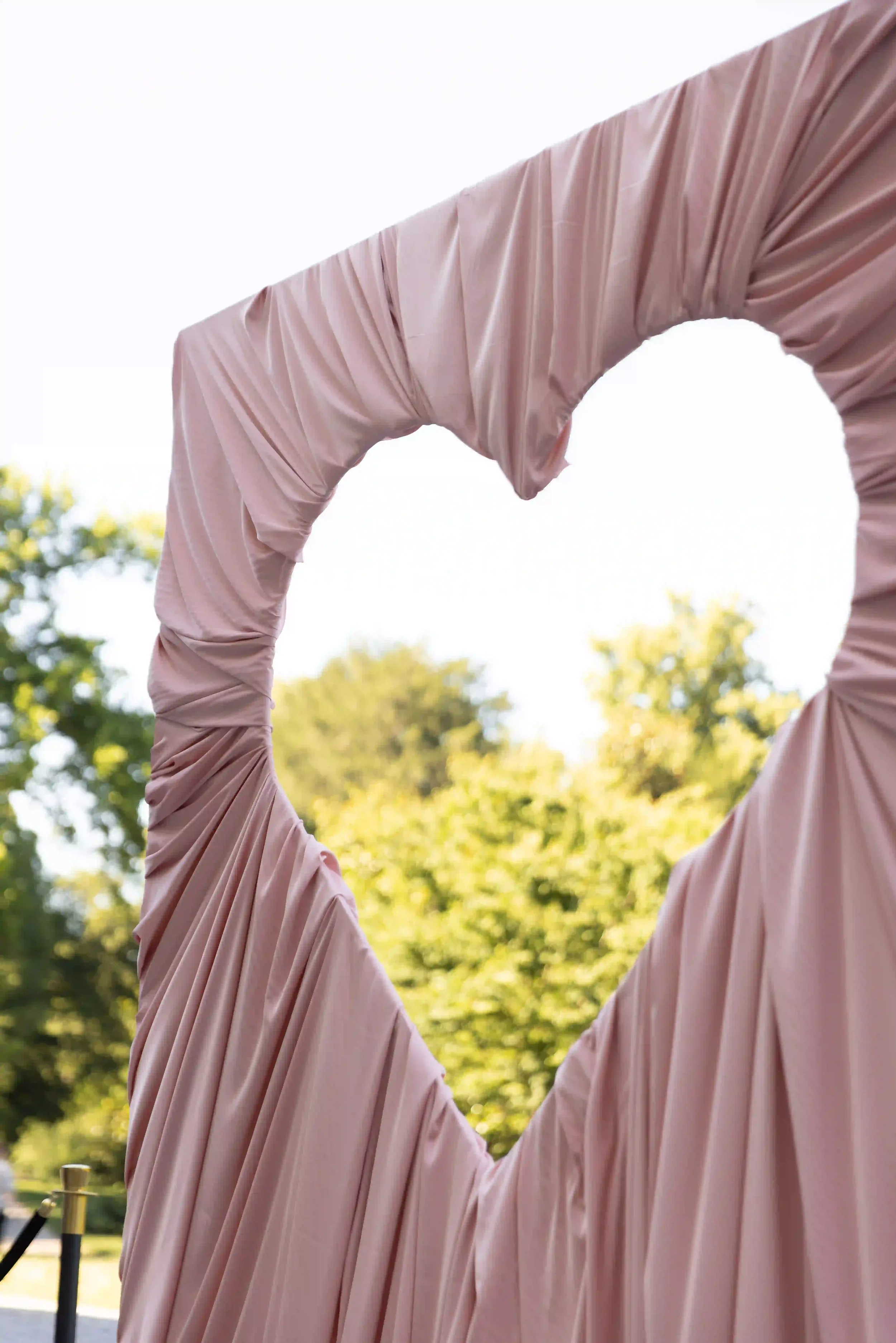 Scenografia outdoor per matrimonio caratterizzata da una grande struttura a forma di cuore interamente drappeggiata in tessuto morbido rosa cipria, posizionata nel parco della villa.