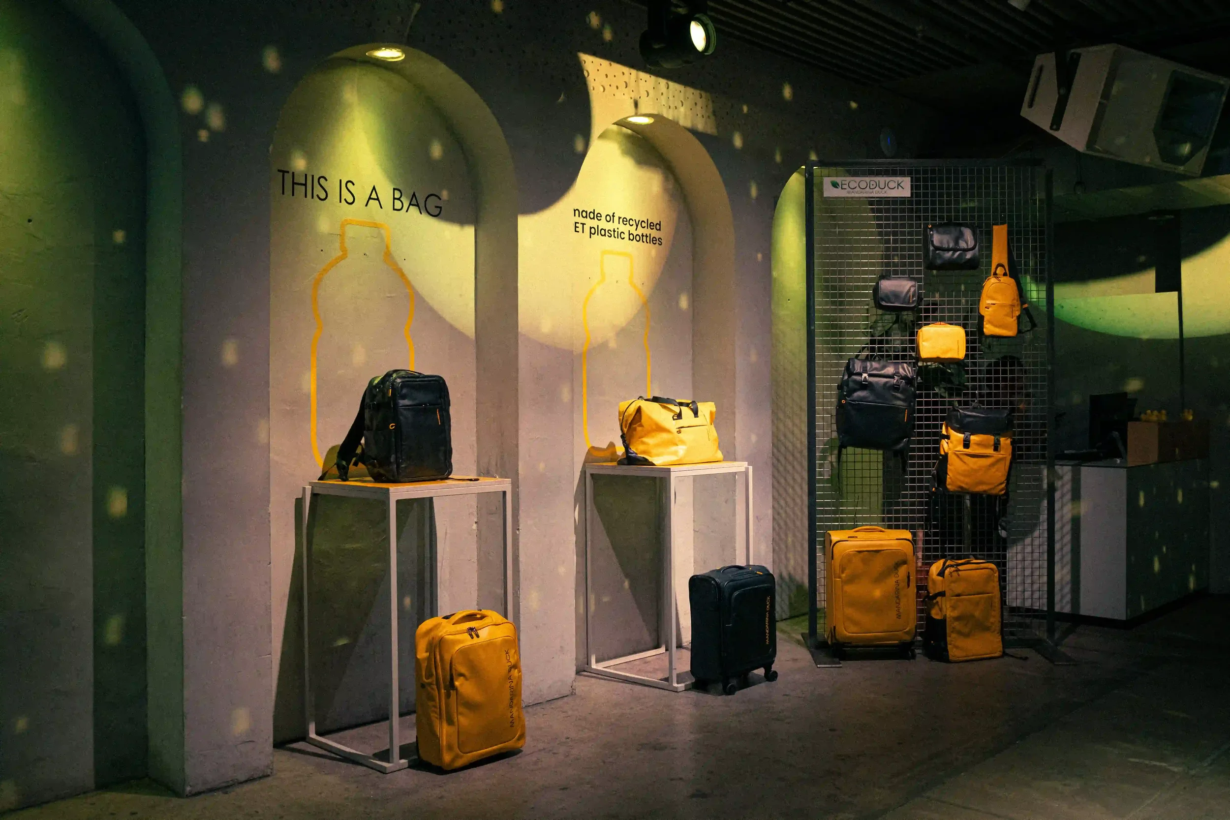 Allestimento espositivo della linea EcoDuck per l'evento Mandarina Duck a Berlino, con zaini e trolley gialli in plastica PET riciclata collocati in nicchie illuminate."