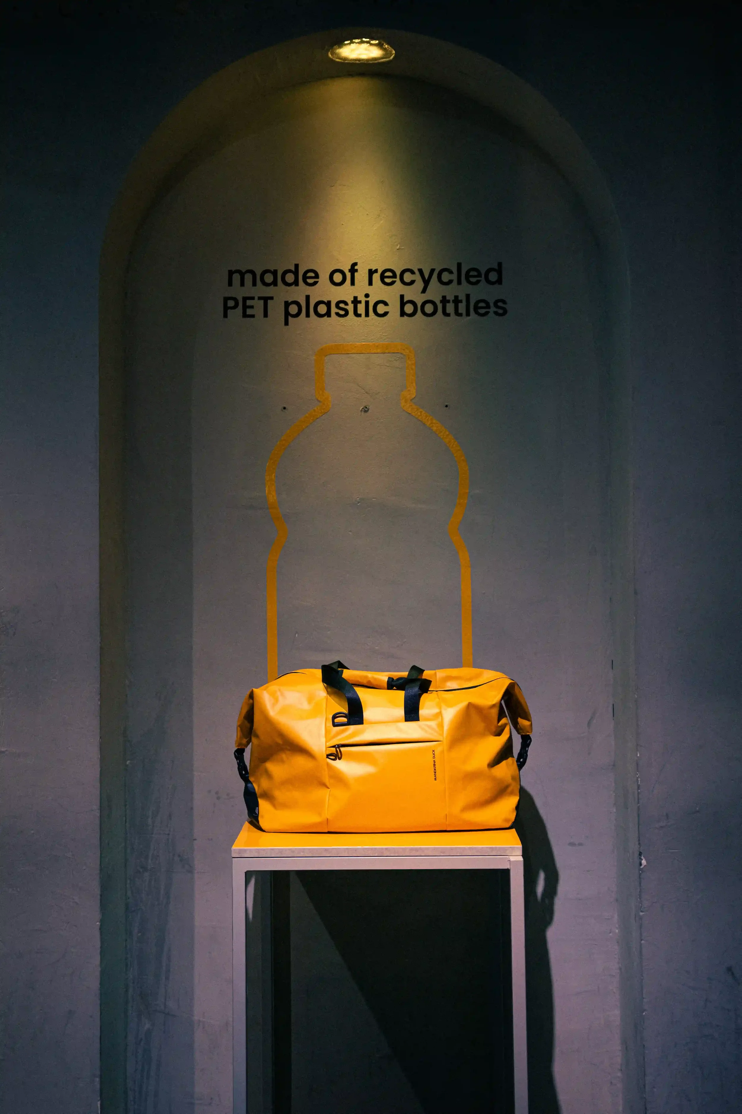 Allestimento espositivo in nicchia con borsone da viaggio giallo Mandarina Duck in plastica PET riciclata, evidenziato da spotlight e grafica a parete all'evento di Berlino.