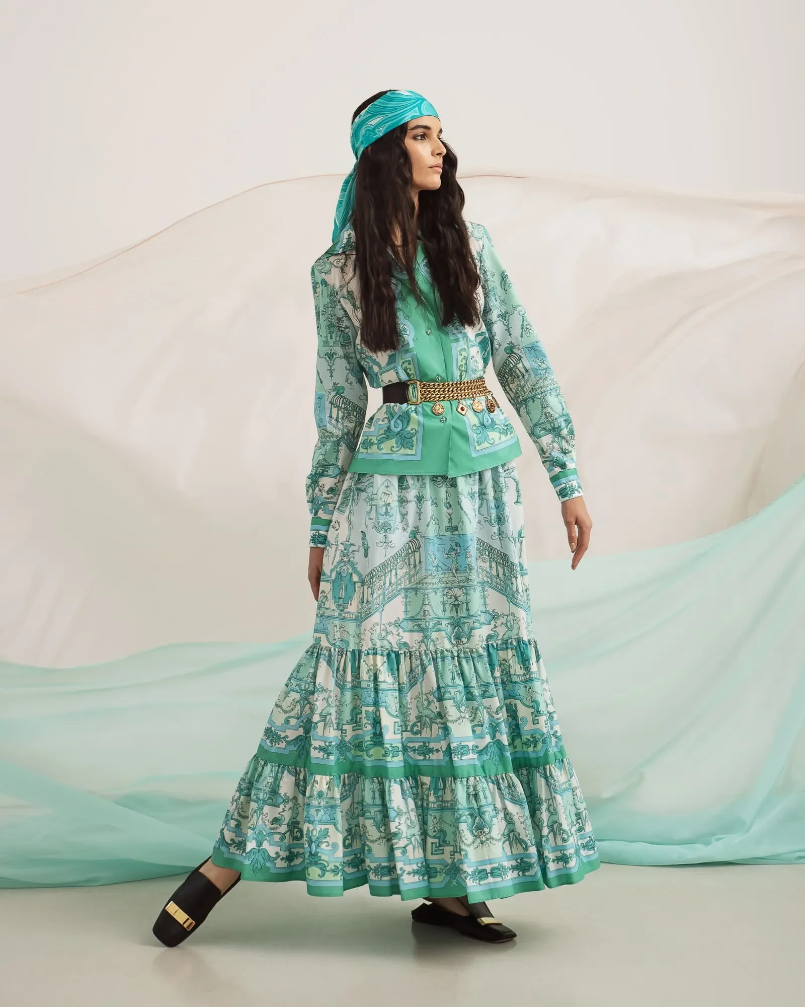 Scatto a figura intera della modella in abito turchese a balze con cintura dorata e foulard, circondata da veli di chiffon verde acqua.