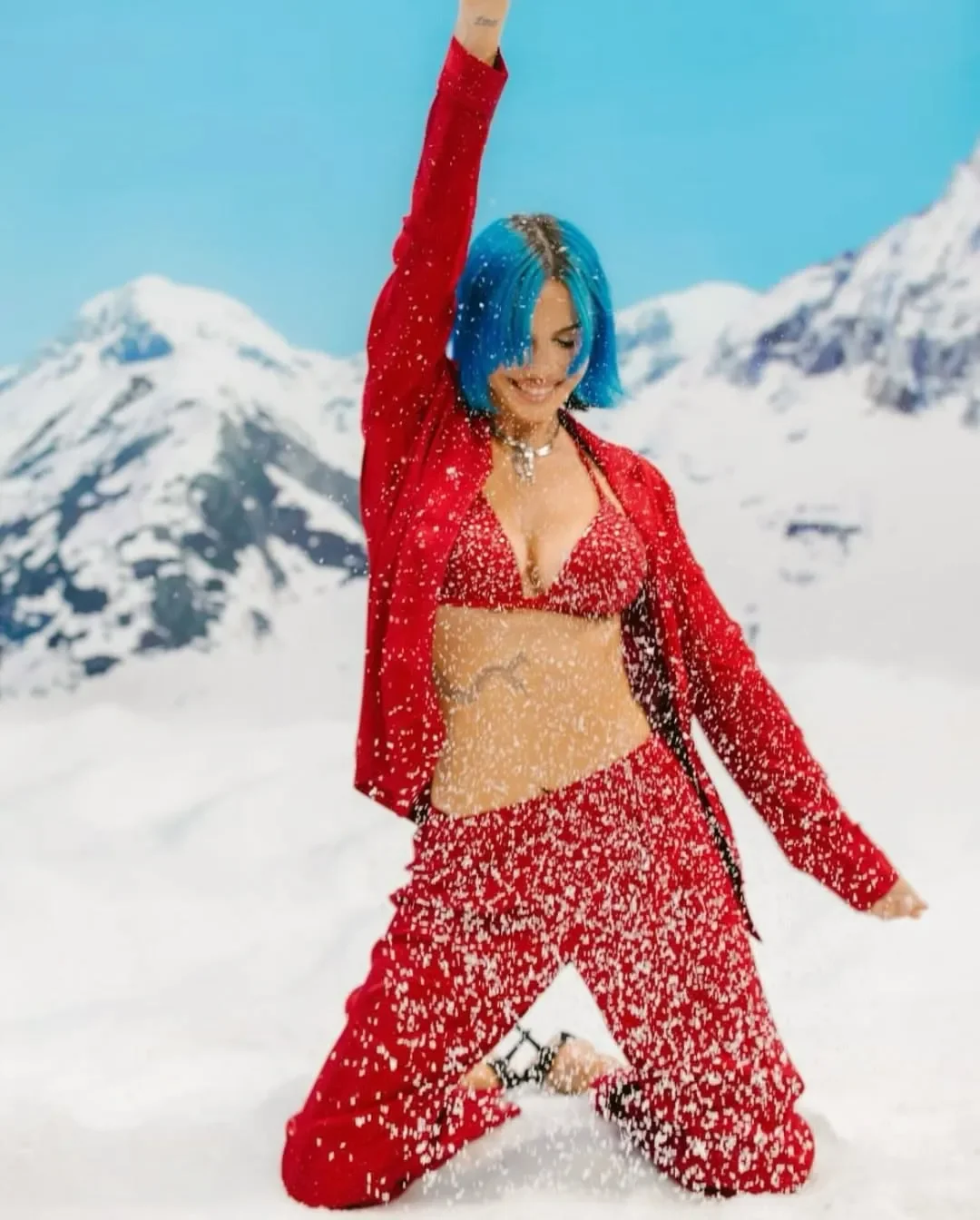 Ritratto in primo piano di Rose Villain accucciata sulla neve con completo pigiama rosso lucido Yamamay e acconciatura blu, in un'atmosfera invernale e natalizia, che gioca con la neve