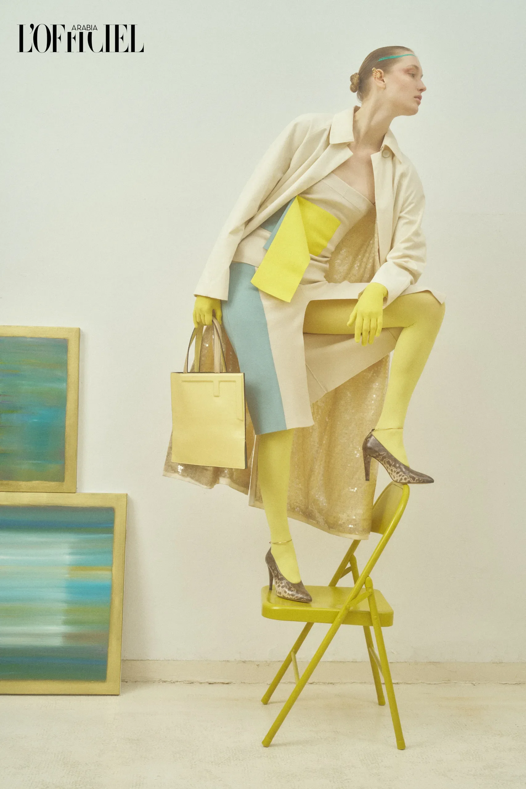 Modella in equilibrio su una sedia giallo limone in uno studio d'arte, con completo sartoriale beige, dettagli color block e borsa coordinata.