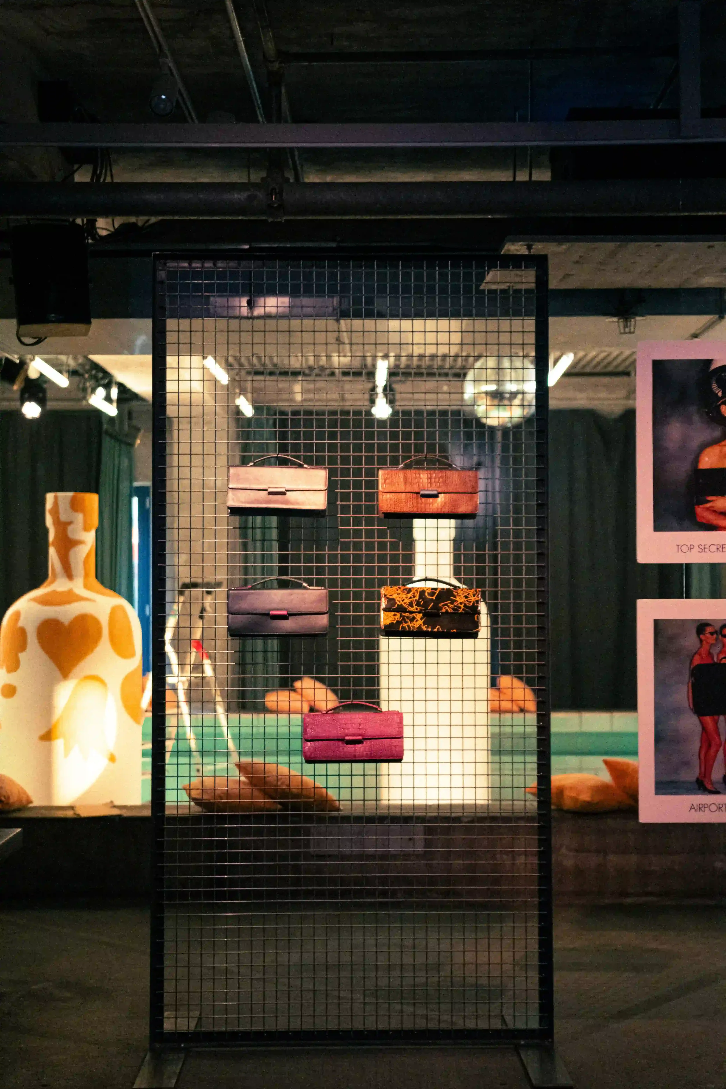 Borse vintage in pelle d'archivio Mandarina Duck presentate su una struttura a griglia in metallo nero, con installazione luminosa a forma di bottiglia sullo sfondo.