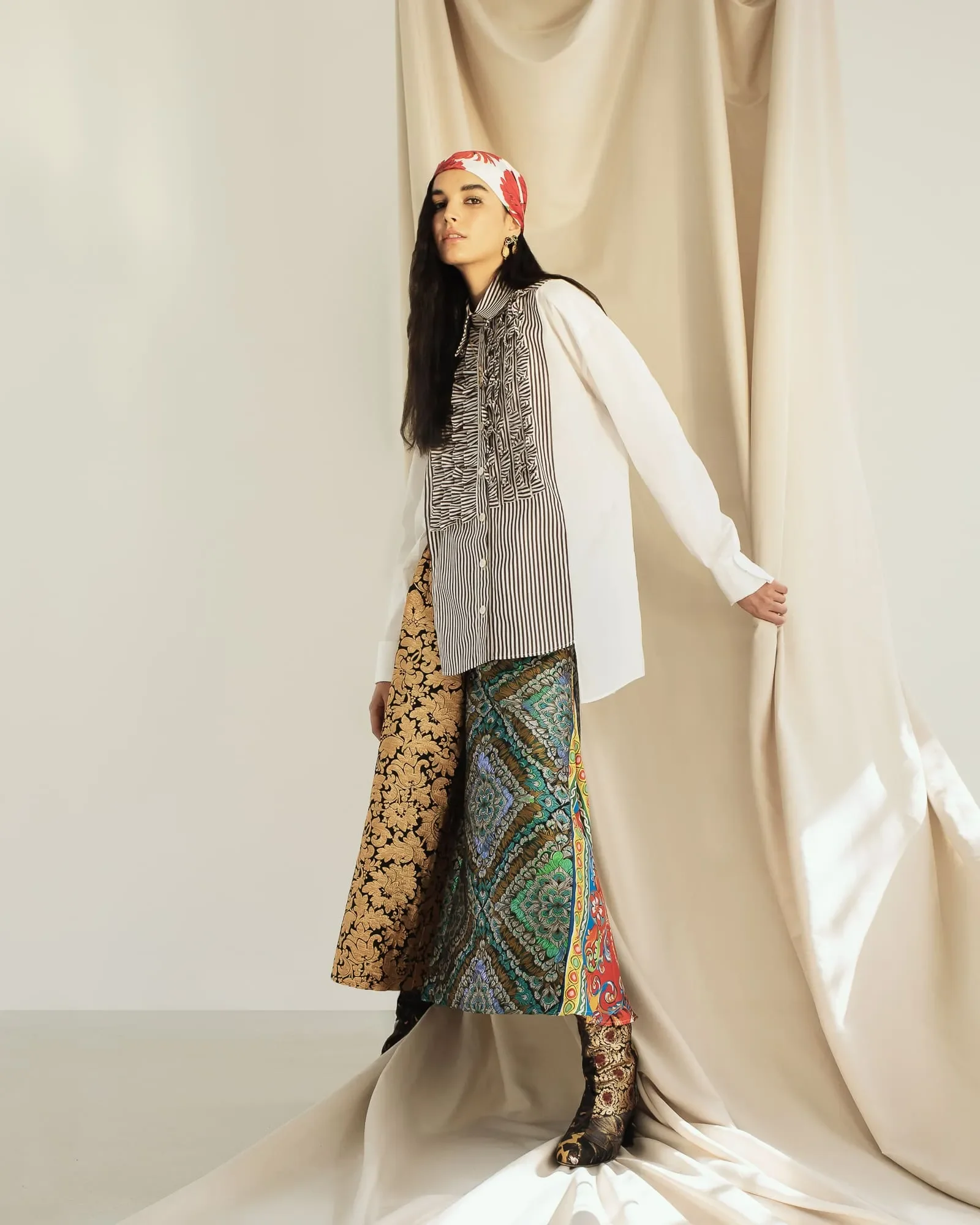 Modella in posa per un editoriale moda con camicia a righe, gonna lunga a motivi e sovrapposizioni geometriche, foulard rosso in testa e sfondo di tendaggi beige."