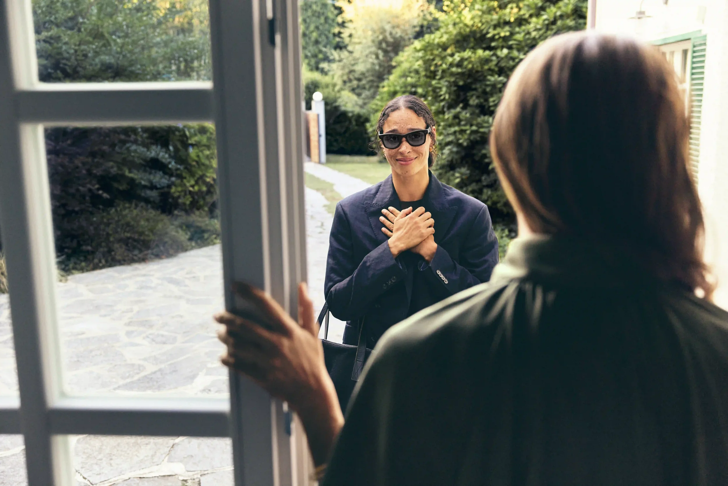 Ritratto lifestyle per il Natale Sunglass Hut con modella all'esterno con le mani sul cuore in un gesto affettuoso verso la padrona di casa, mentre indossa occhiali da sole neri eleganti.