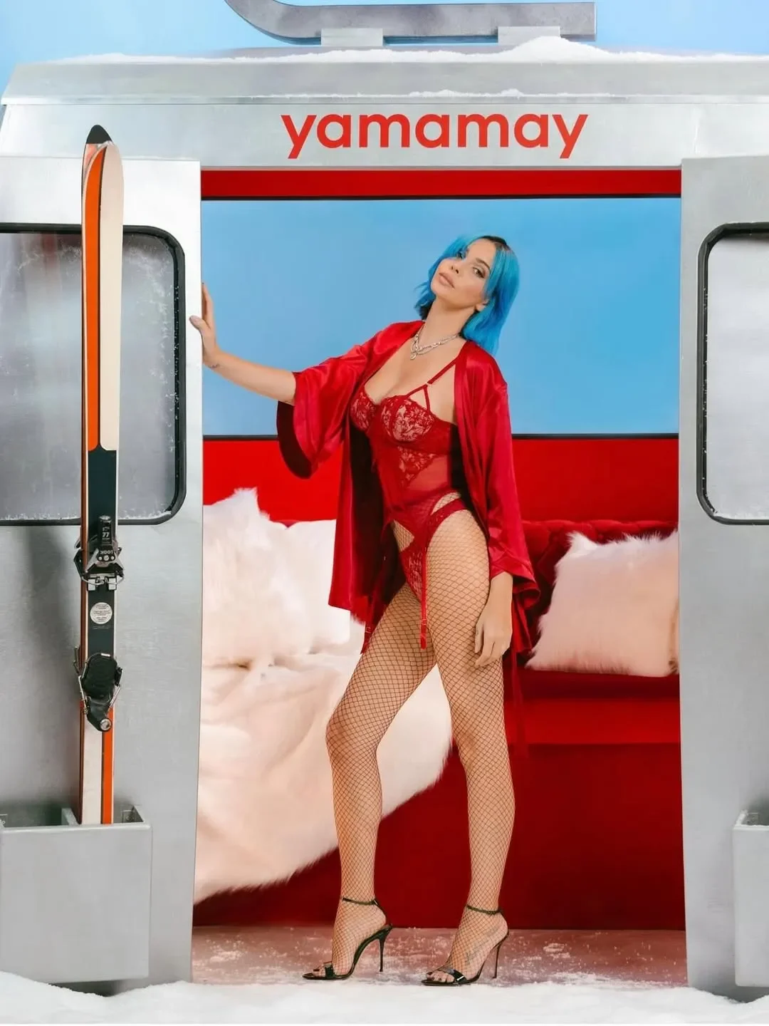 Scatto pubblicitario della campagna natalizia Yamamay con Rose Villain dai capelli blu in lingerie di pizzo rosso e vestaglia in raso, in posa sulla porta di una cabinovia vintage da sci