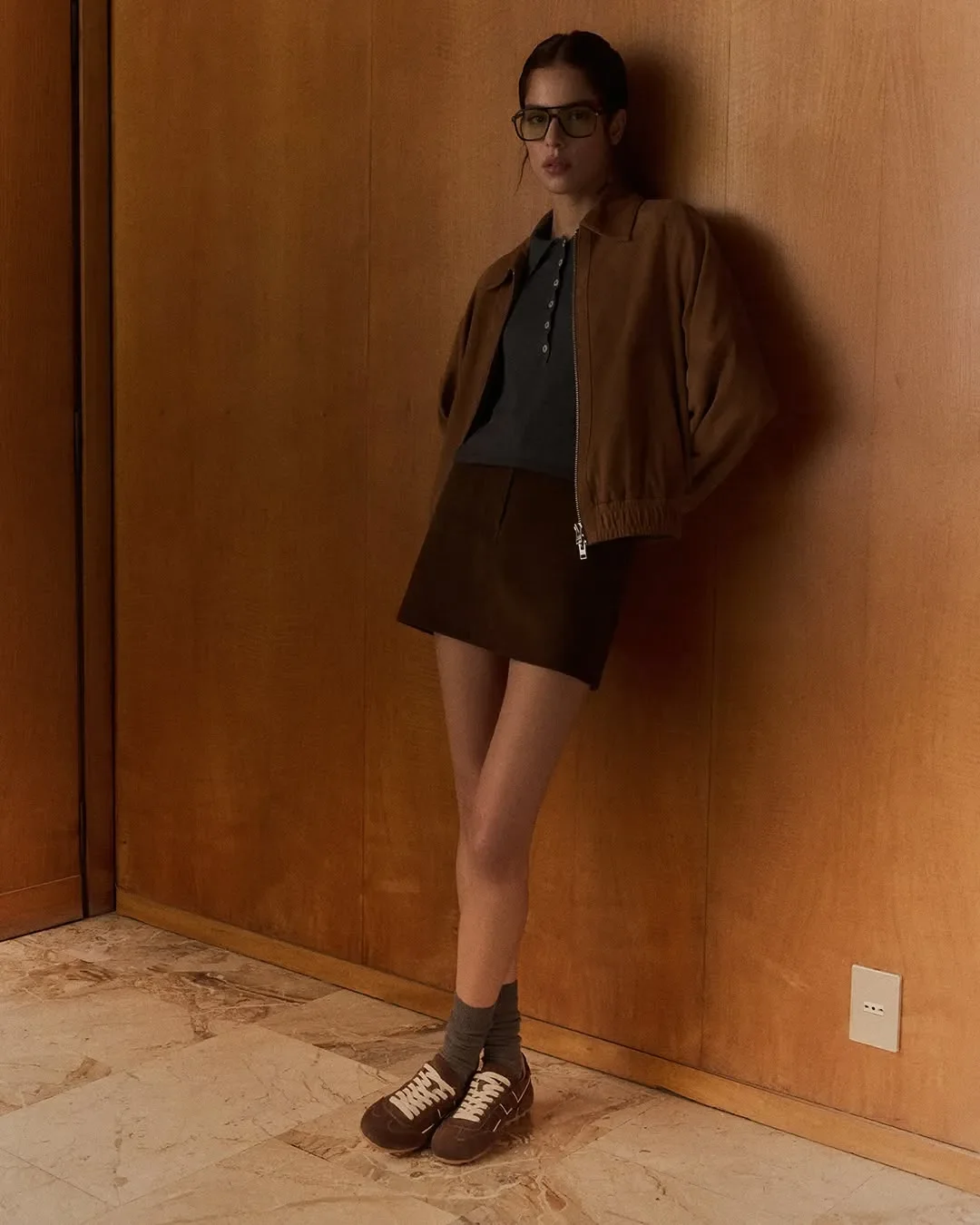 Scenografia d'interni e ritratto editoriale per la campagna Hogan. Modella con giacca in camoscio, occhiali da vista e sneakers marroni appoggiata a una parete in legno vintage.